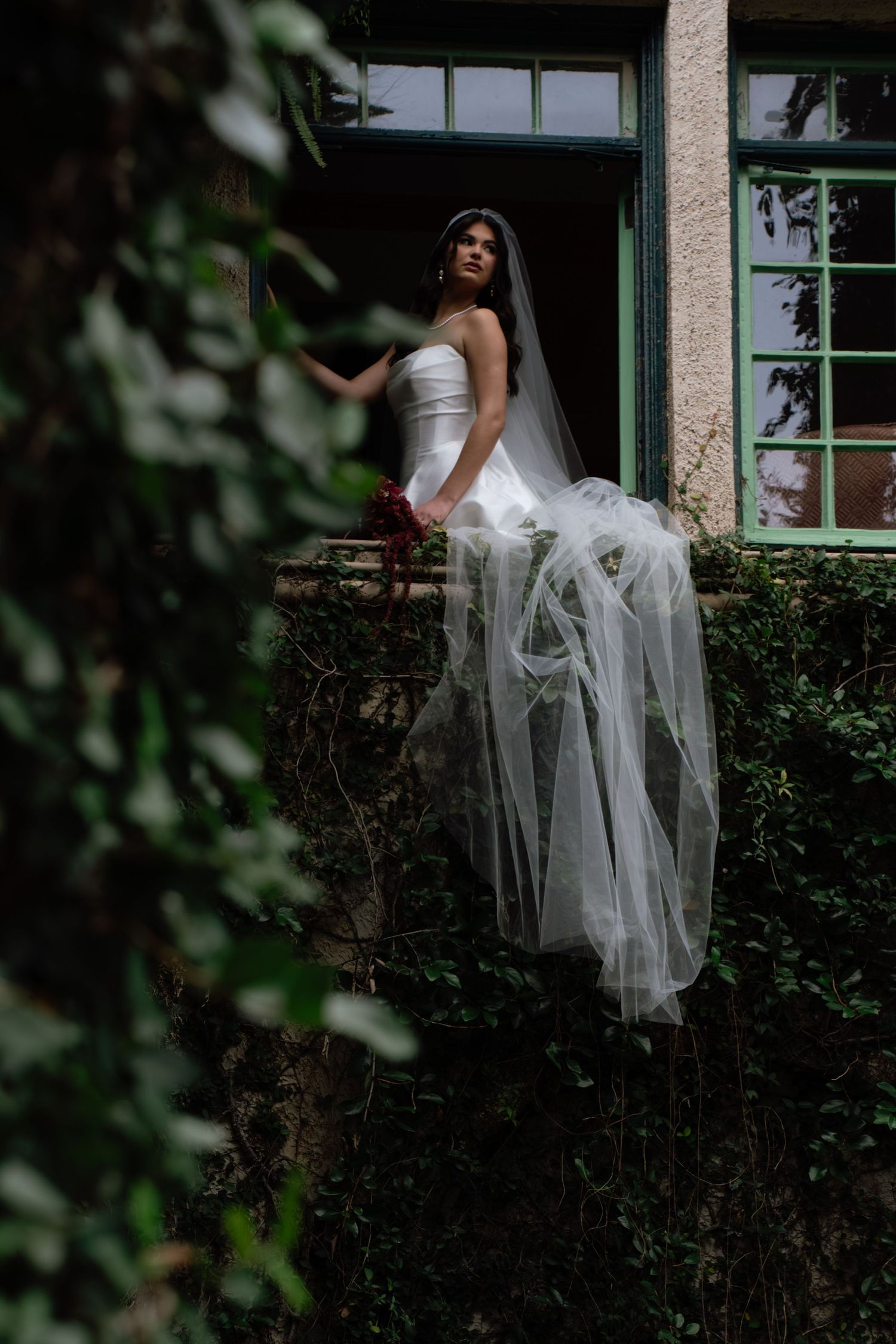 A bride in a white dress and flowing veil poses gracefully against a stone wall with ivy and windows in the background.