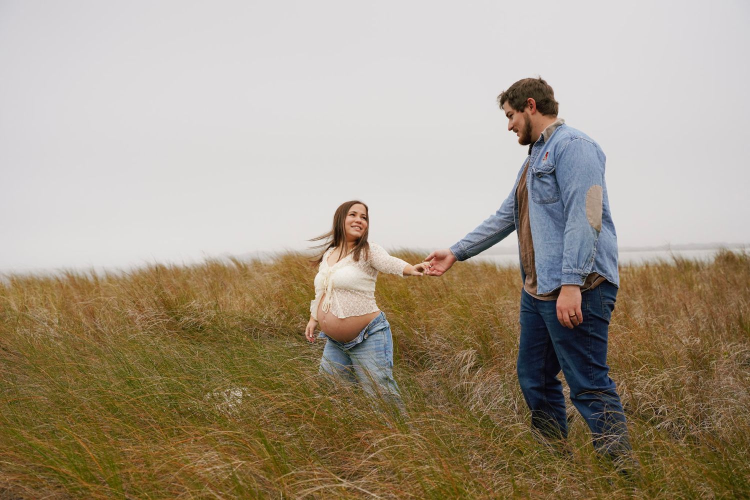 A couple walks hand in hand through a serene golden field on a cloudy autumn day.
