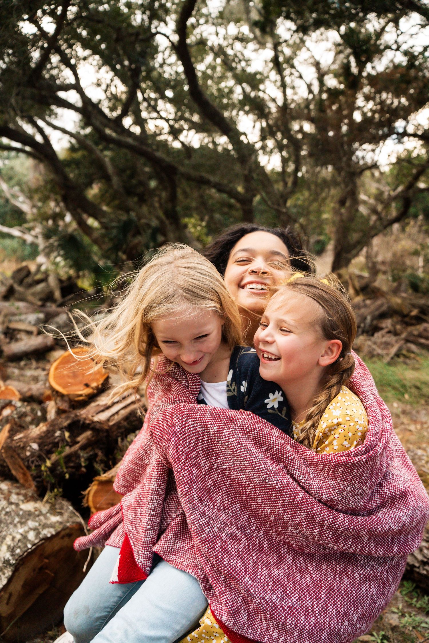 Three children laughing and hugging while wrapped in a pink blanket outdoors.