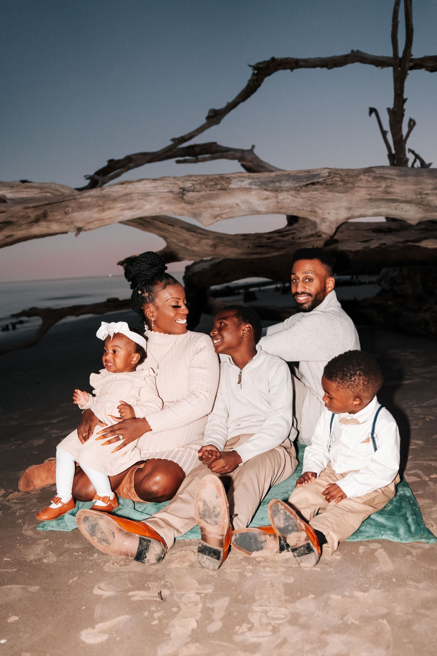 Family in white outfits sitting together on beach at sunset near driftwood.