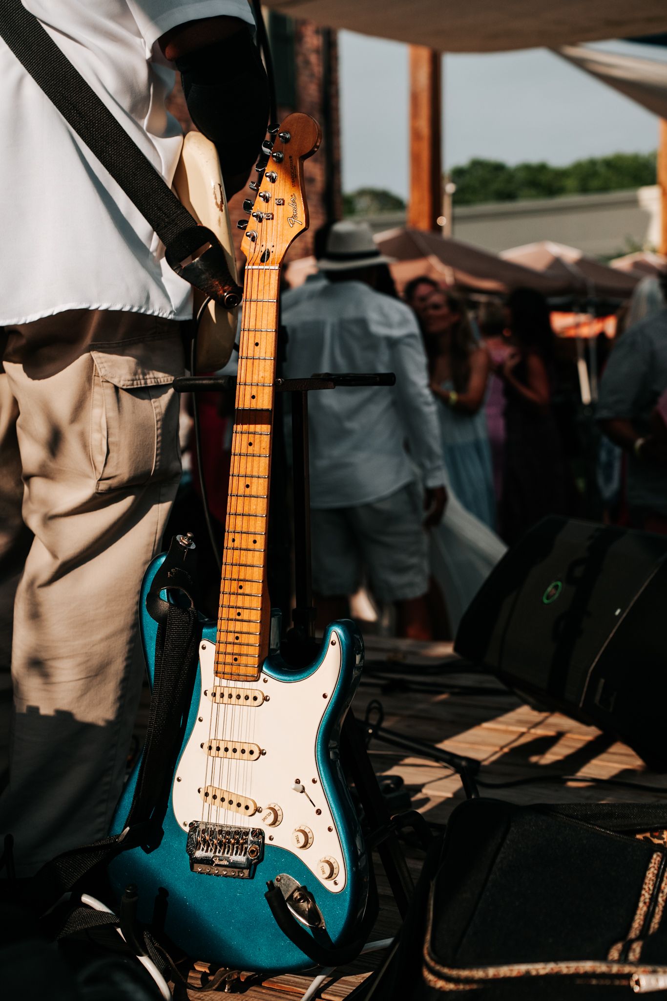 A teal Fender Stratocaster electric guitar resting on stage at an outdoor music event in warm sunlight.