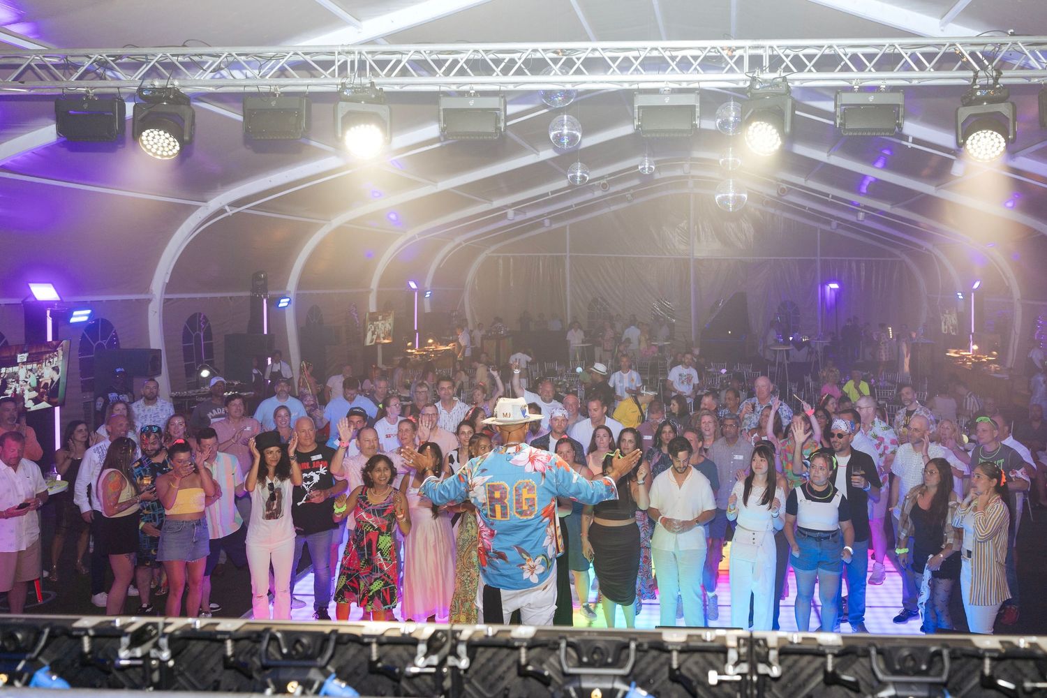 Large crowd gathered under a tent at a vibrant outdoor party event with colorful stage lighting and performers.