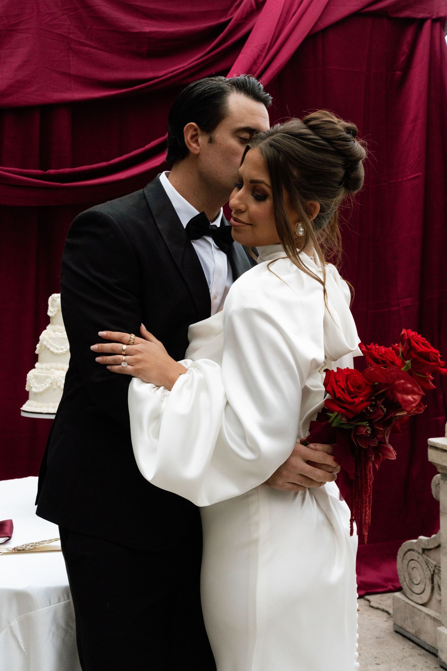 Romantic wedding couple embraces against burgundy drapes, bride in white dress holding red roses.