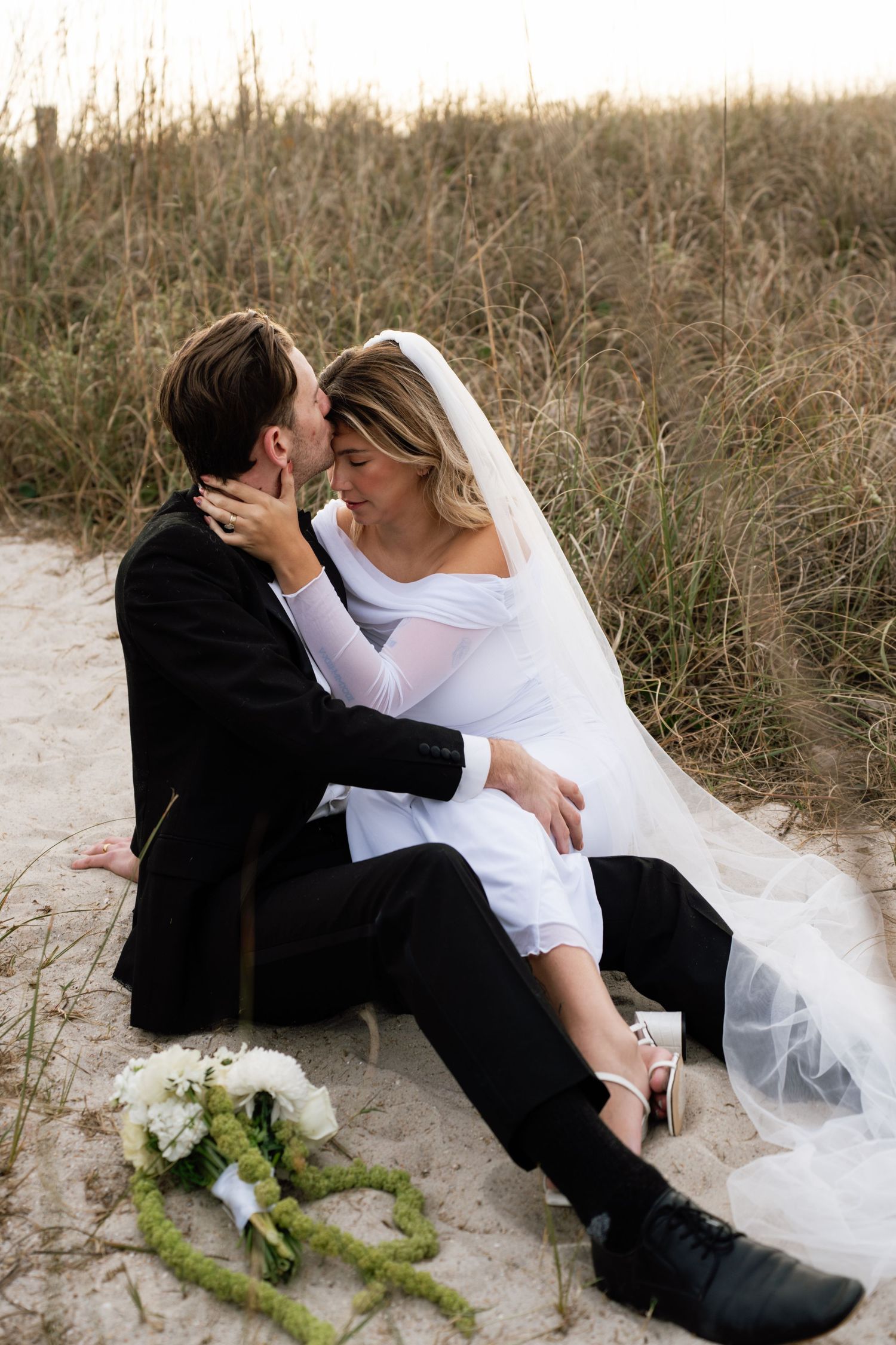A romantic beach wedding photo series shows a couple in formal attire reclining in the sand dunes at sunset.