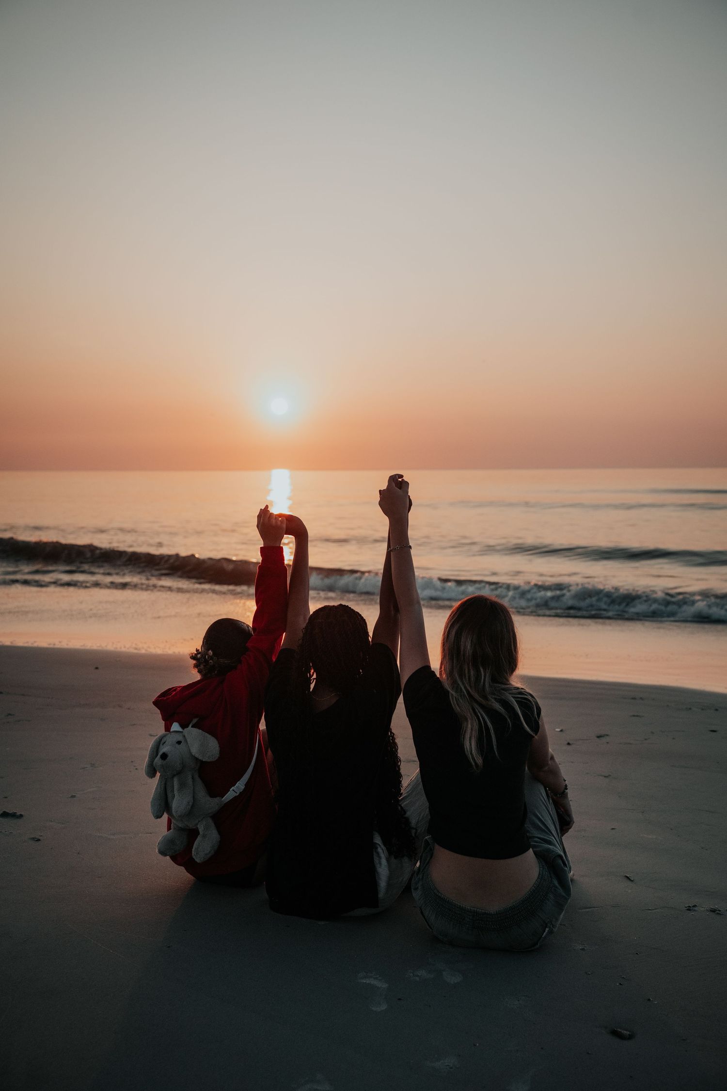 Friends sitting on the beach at sunset raising their arms in celebration silhouetted against an orange sky on Amelia Island in Fernandina
