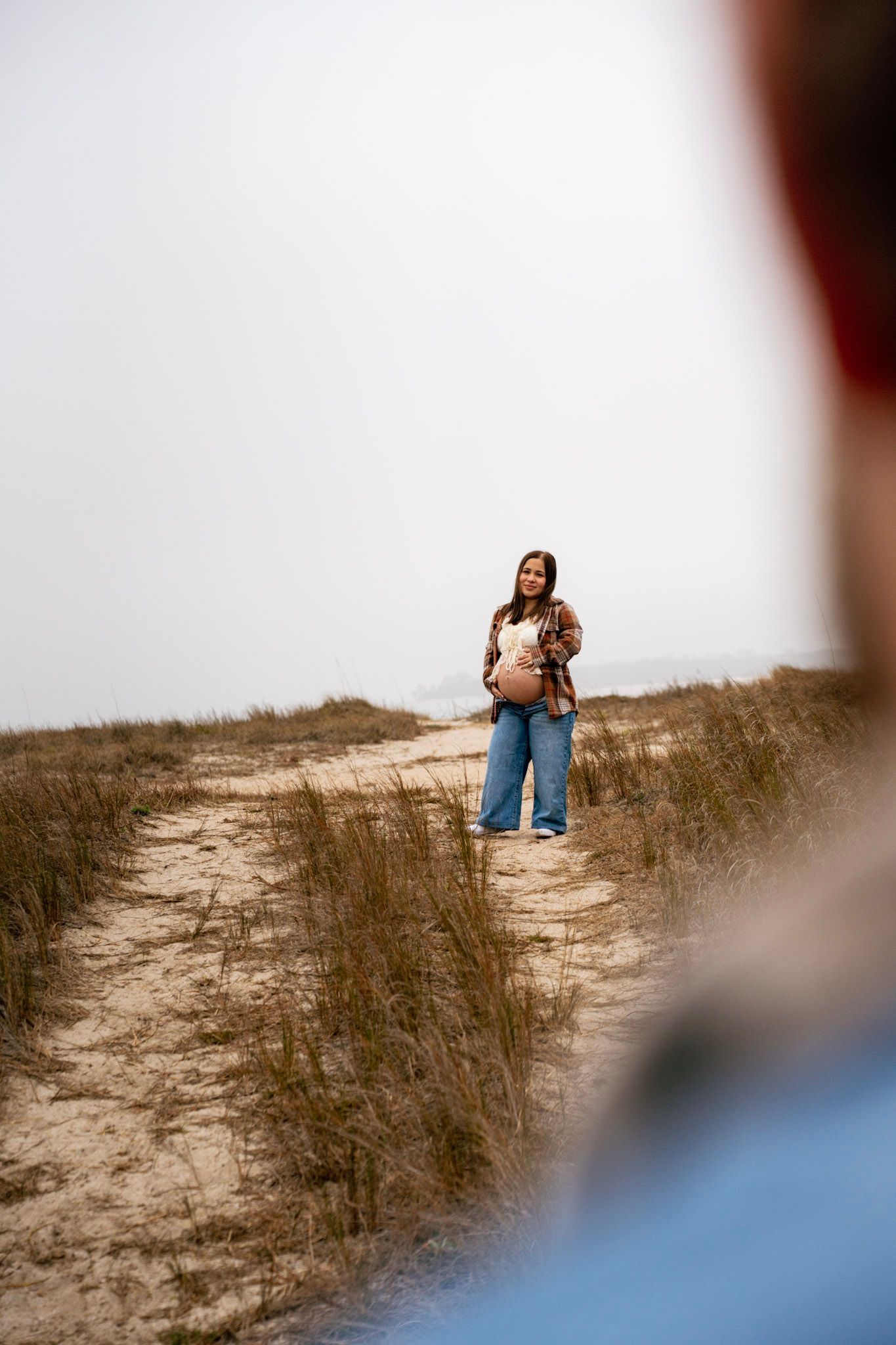 A person in jeans sits on a sandy dune hill during sunset, with a blurred foreground element.