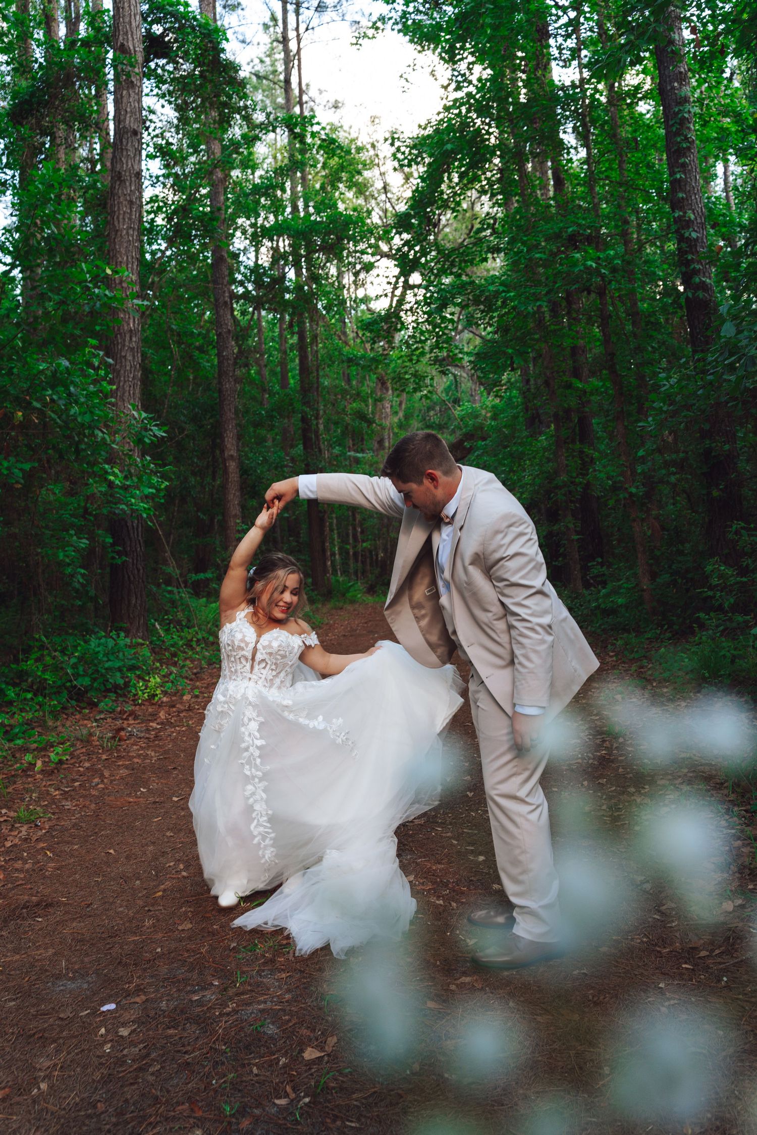 A romantic wedding photoshoot in a lush green forest shows a couple dancing together on a dirt path.