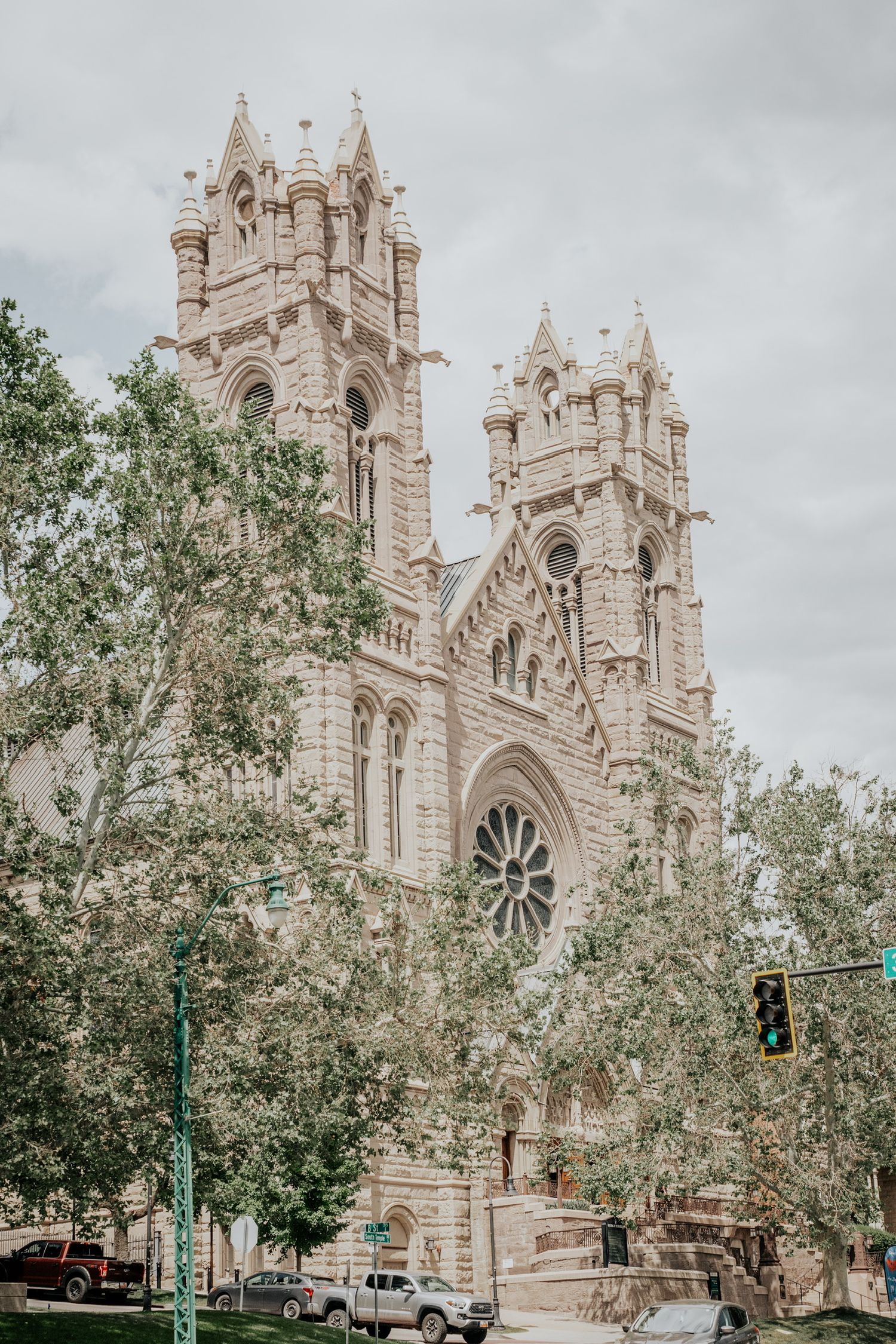 Gothic cathedral with ornate stone spires rises behind green trees against cloudy sky in Salt Lake City.