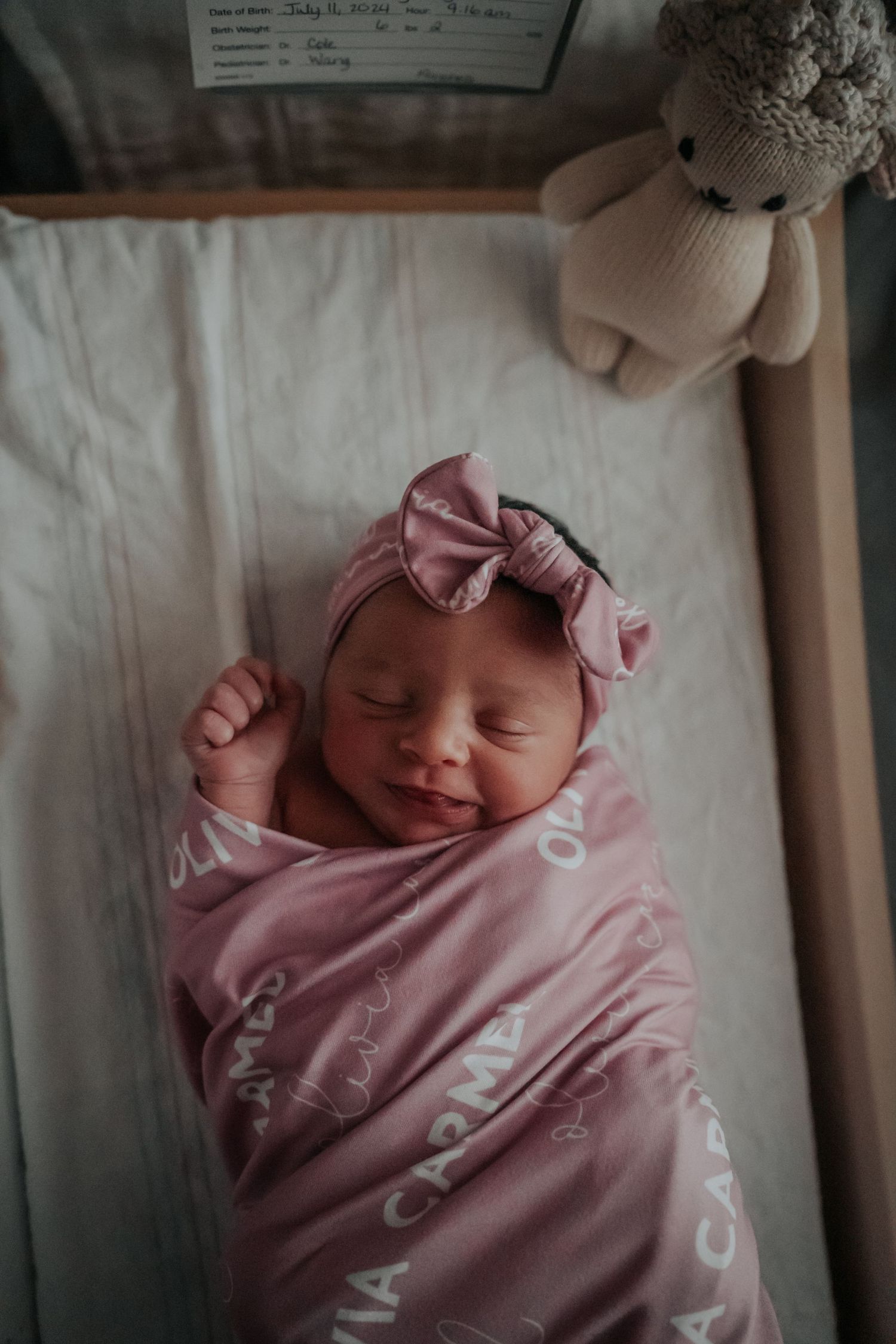 Sleeping newborn wrapped in pink blanket with bow headband next to stuffed teddy bear.