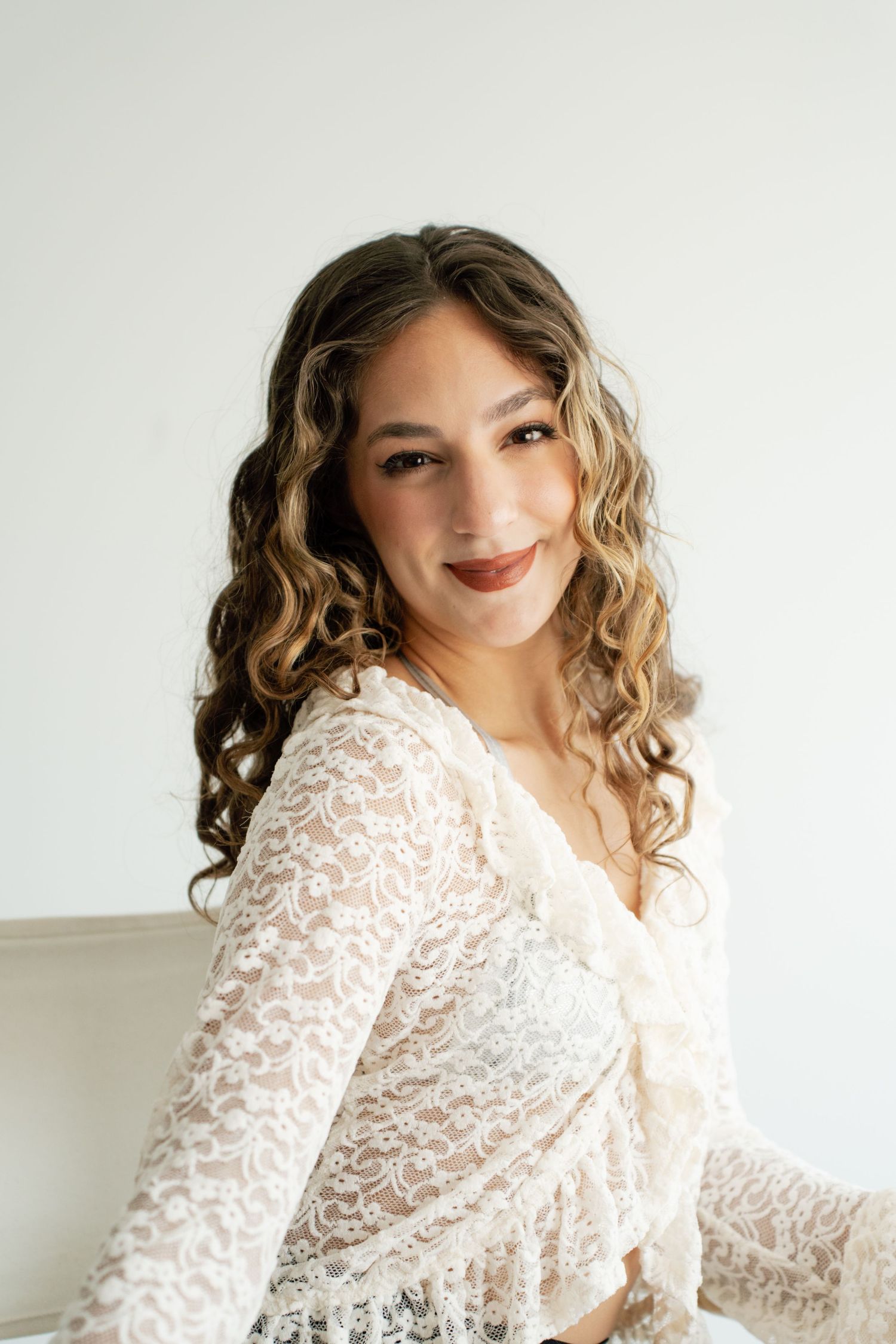 Smiling bride with curly hair wearing a white lace top poses against a white background.