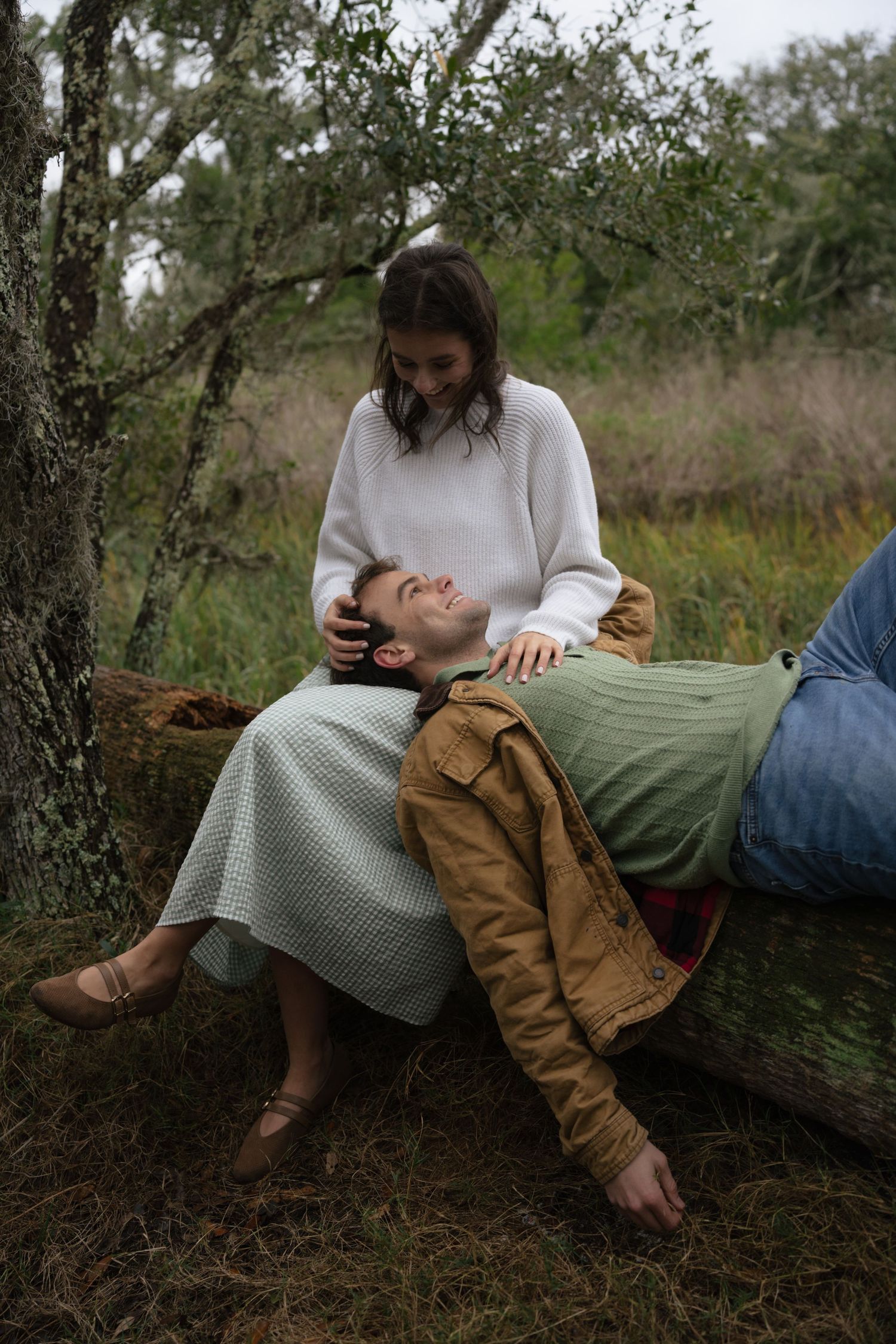 A couple lounges on a fallen log in a serene woodland setting during autumn.
