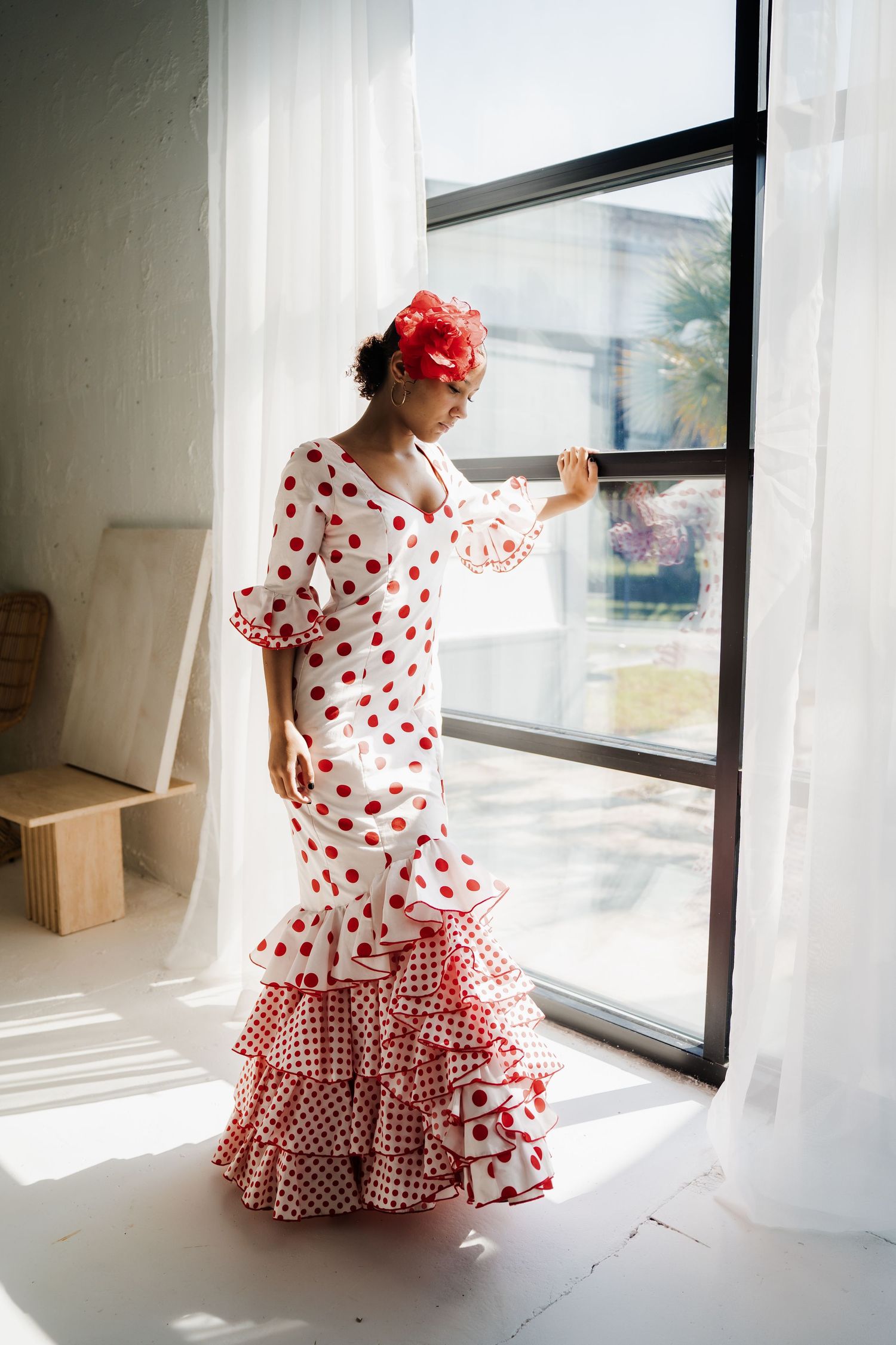 Traditional Spanish flamenco dress with red polka dots and ruffled hem shown near sunny window in white room.