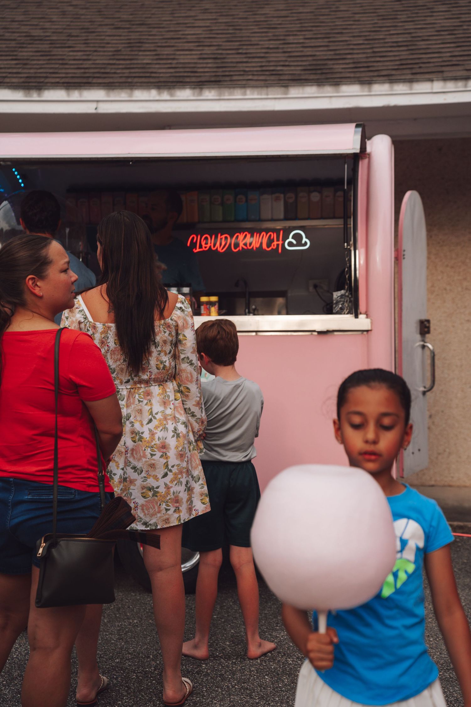 Children and adults queue at a pink Cloud Crunch cotton candy food truck, with a boy holding pink cotton candy in foreground.