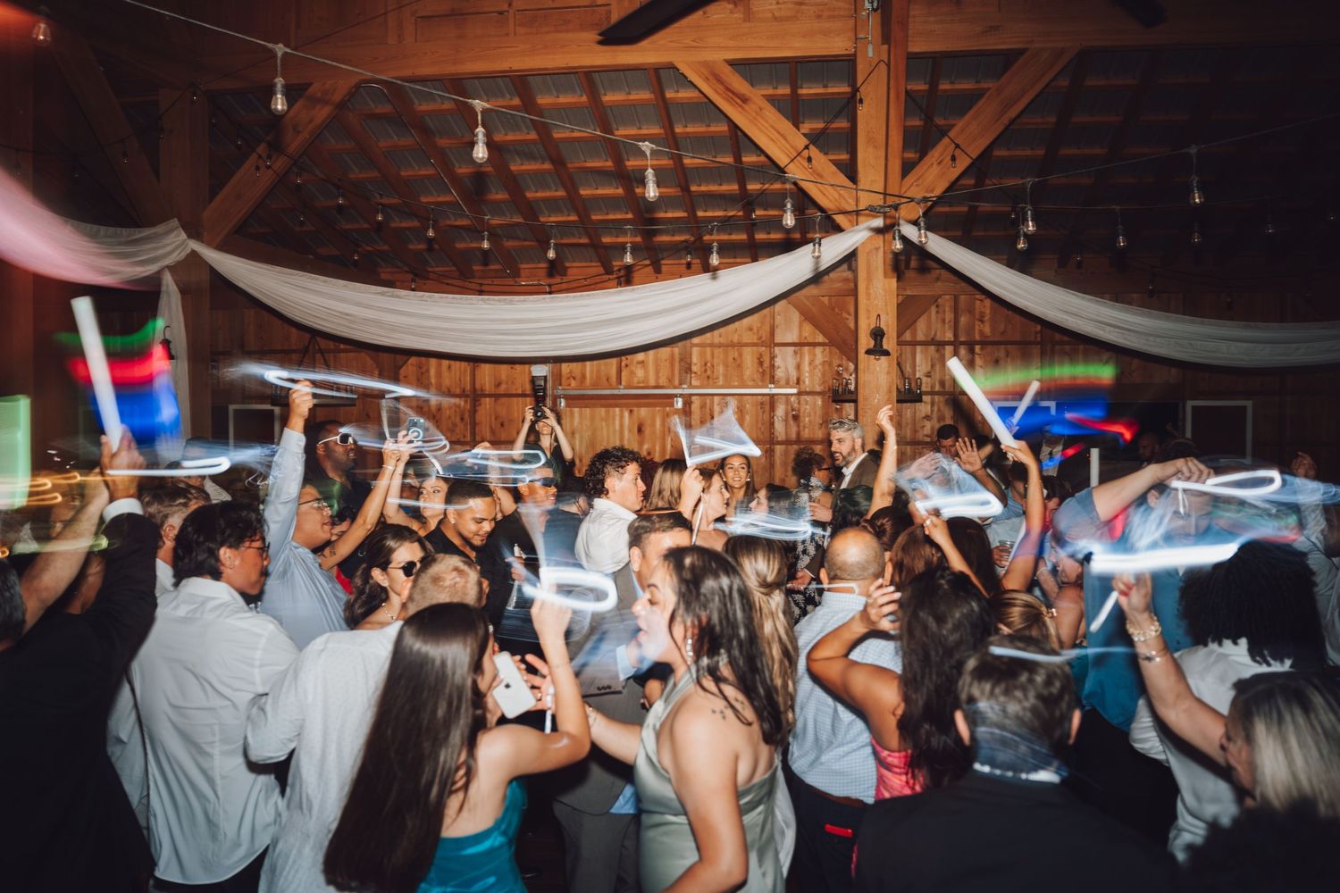 Guests dancing and celebrating at a rustic barn wedding reception with glowing light sticks.