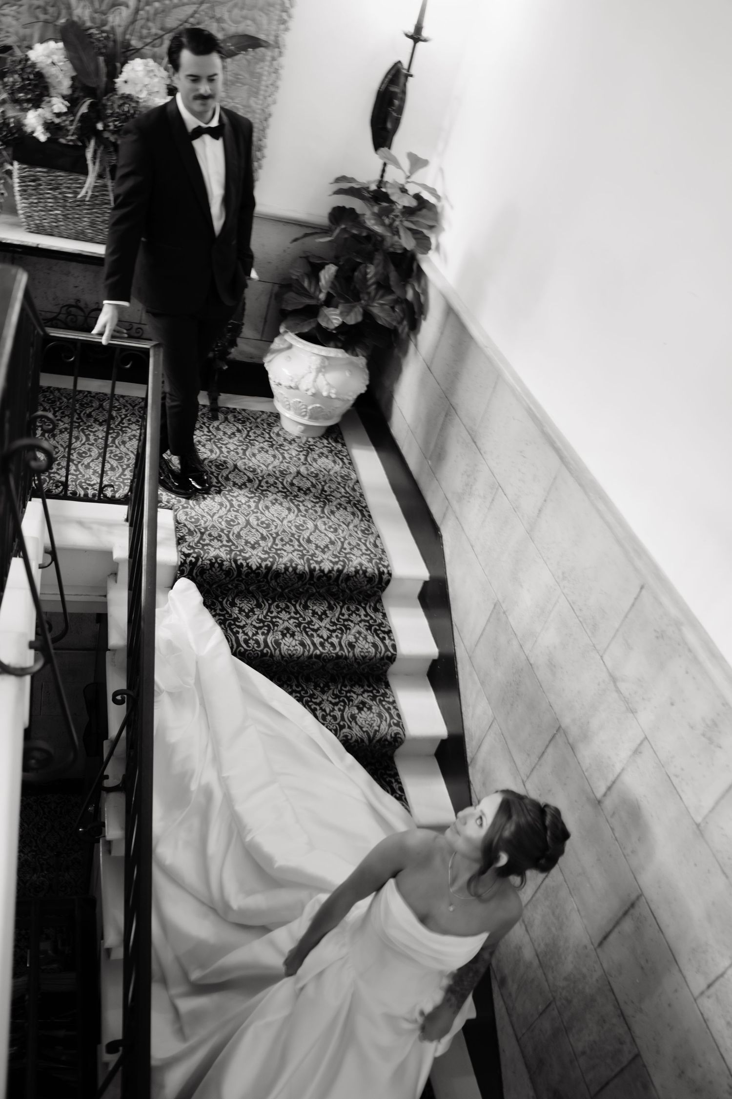 A black and white wedding photograph showing the bride's white dress flowing down a staircase with elegant railing details.