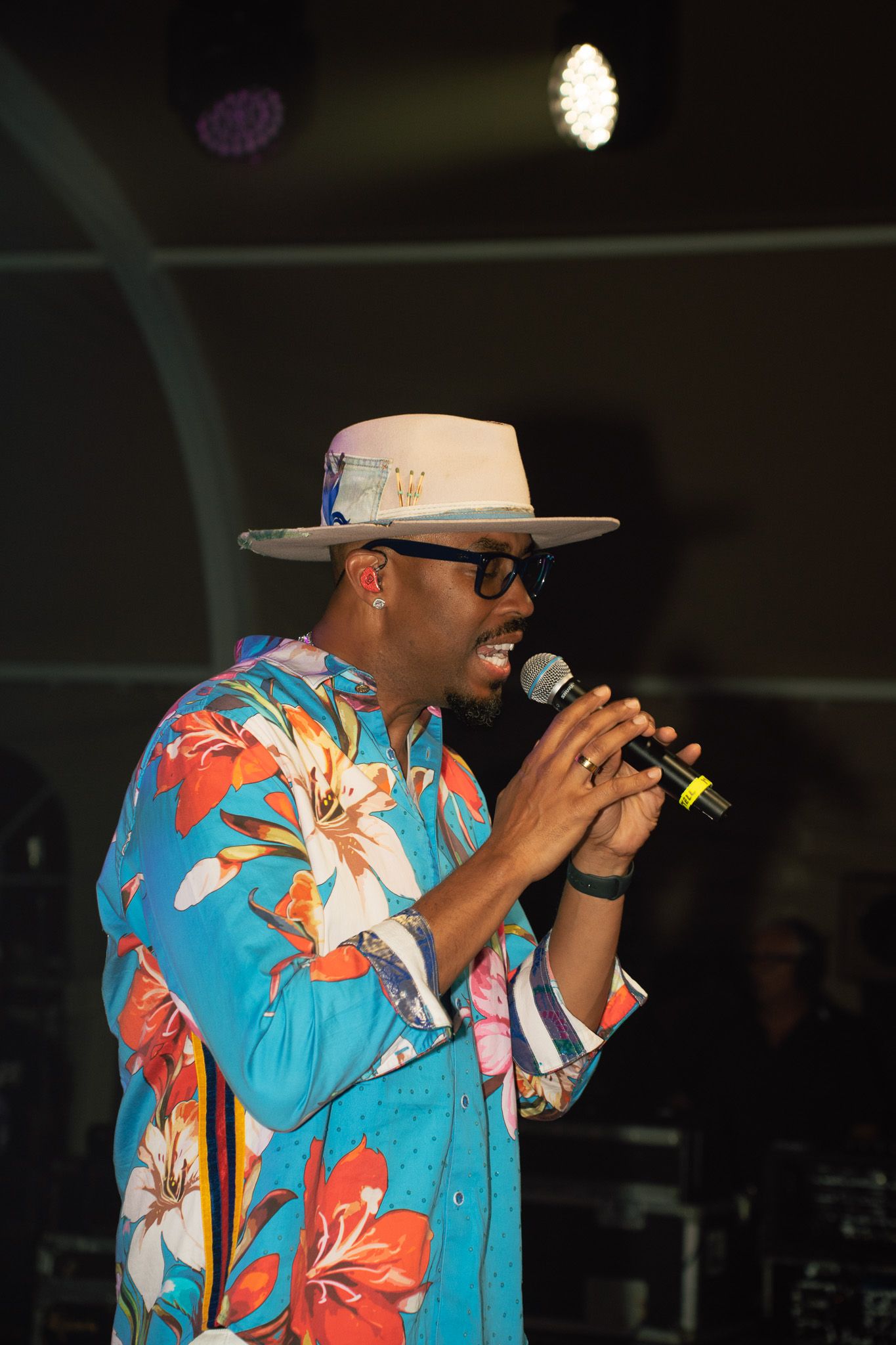 Man in floral shirt and white hat singing into microphone on stage under bright lights.