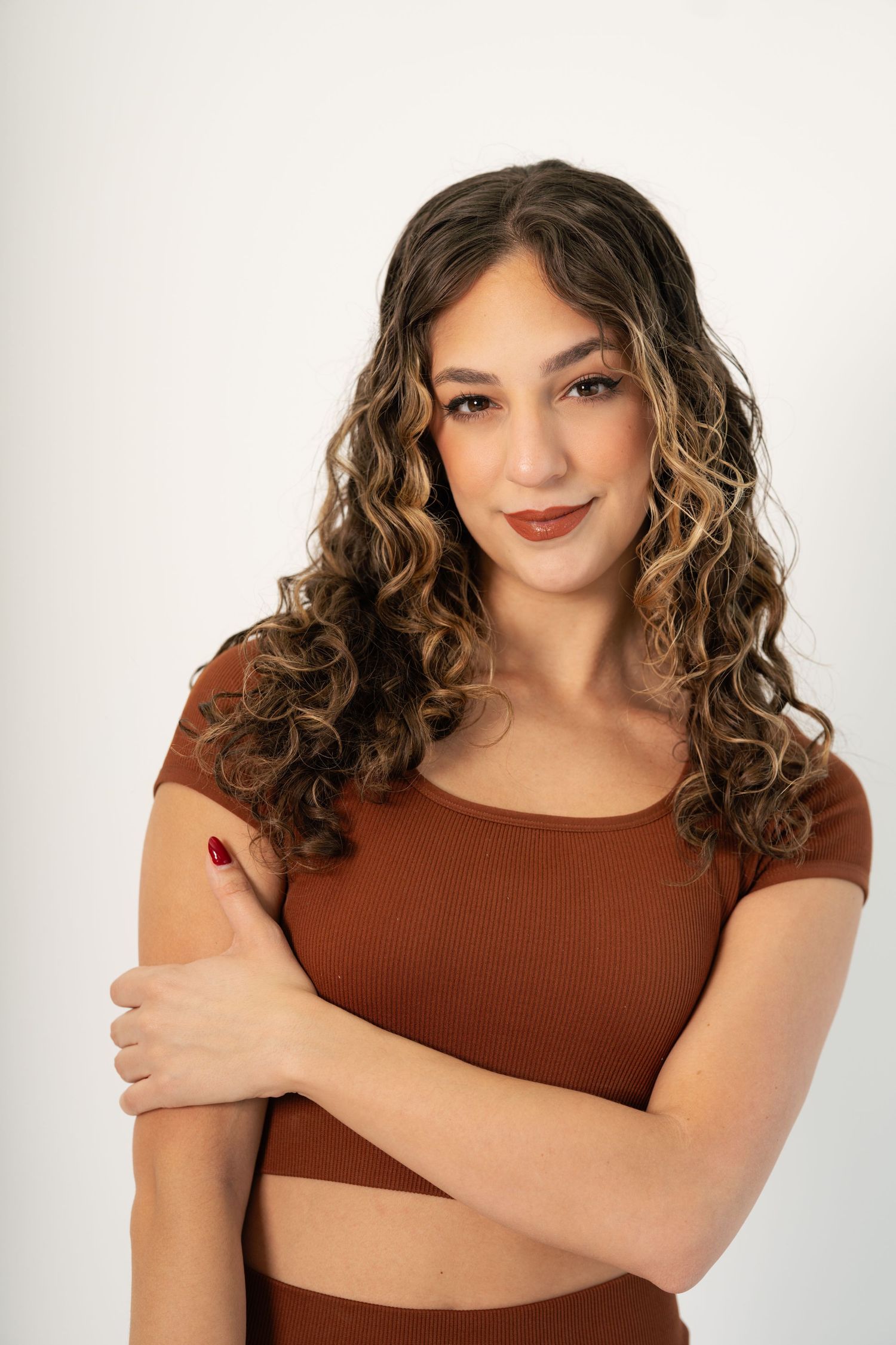 Young woman with curly highlighted hair wearing a brown crop top, arms crossed, smiling against white background.