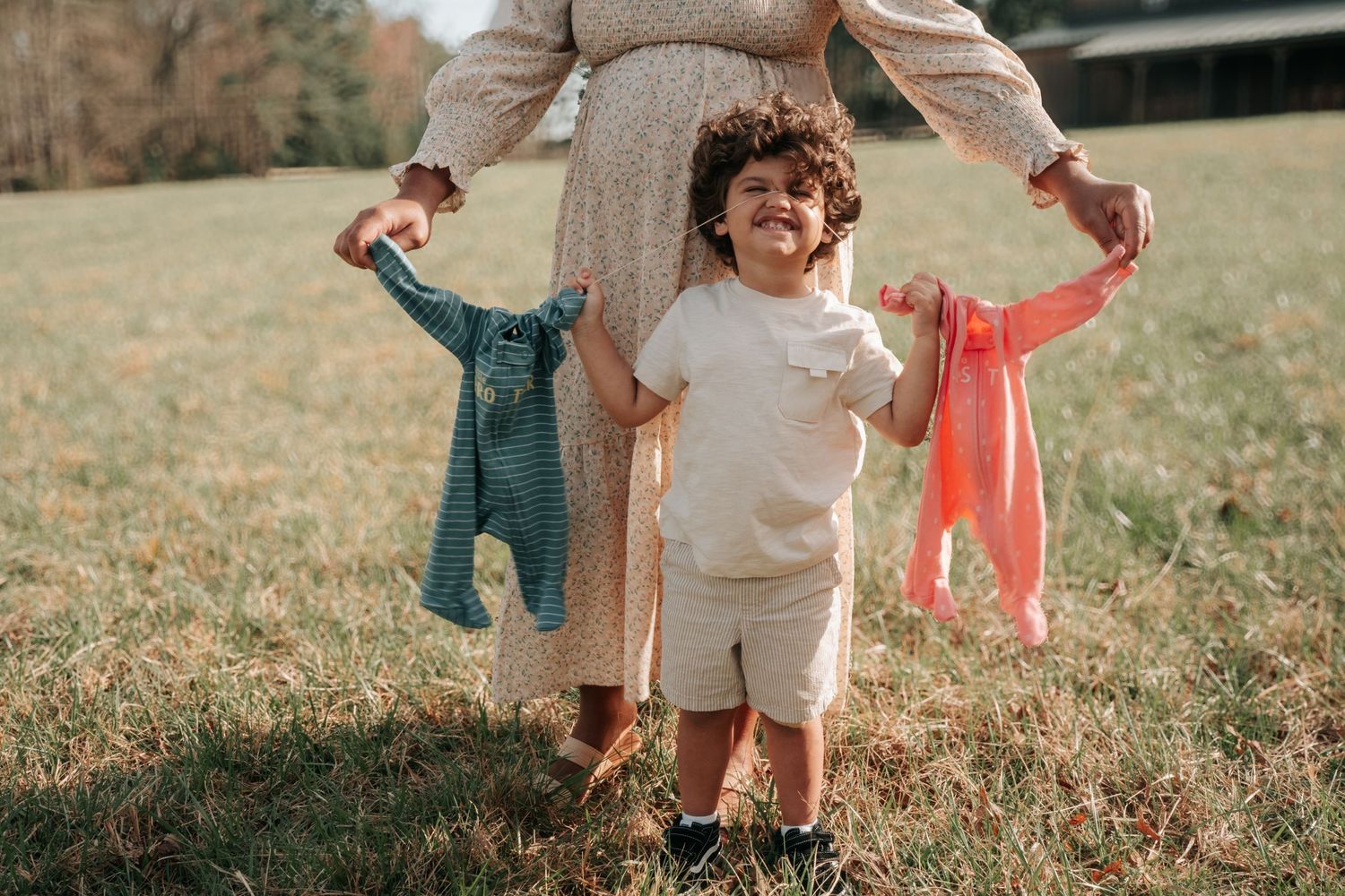 Big Brother stands in a grassy field of Carlee Farms while Mom holds up two colorful baby outfits on hangers for a Maternity Session in NC
