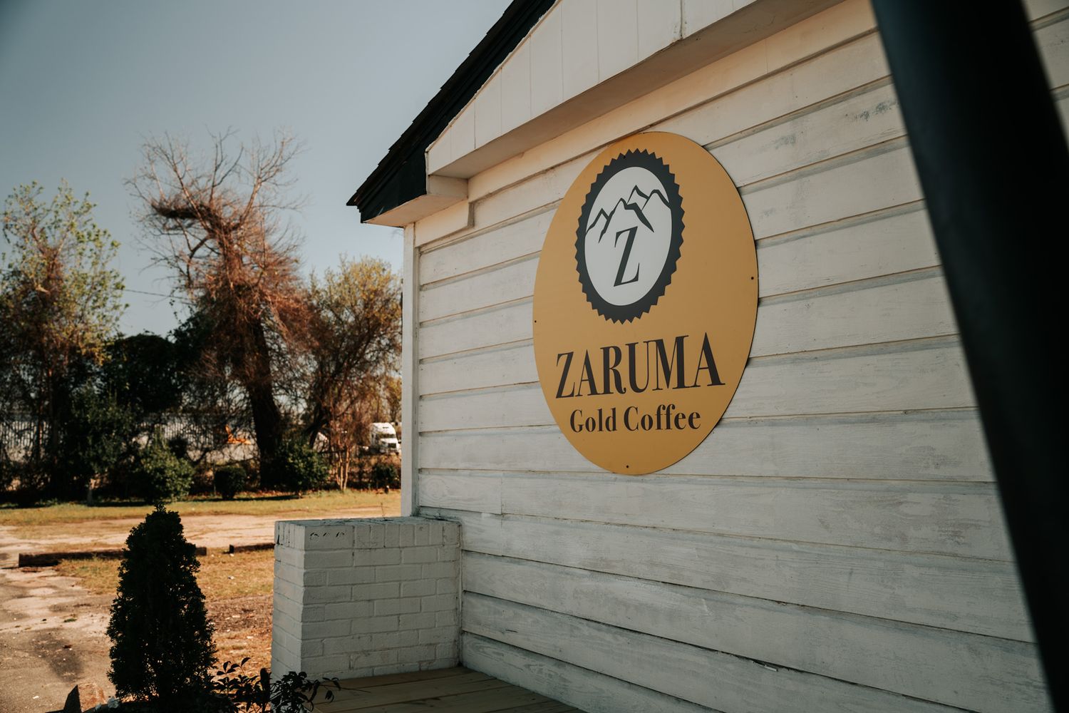 A rustic white building with the Zaruma Gold Coffee logo and text displayed on a circular wooden sign.