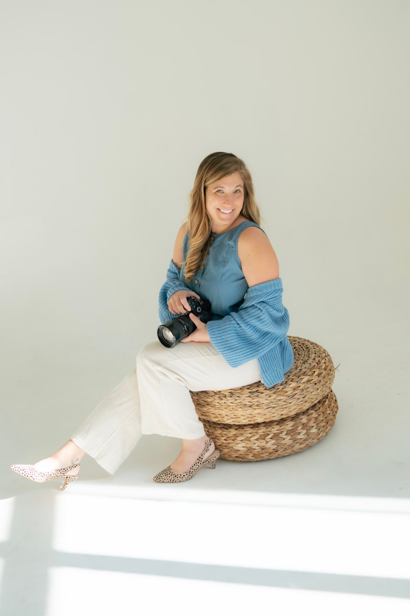 A person in a blue top and white pants lounges on a round woven ottoman in a bright studio setting.