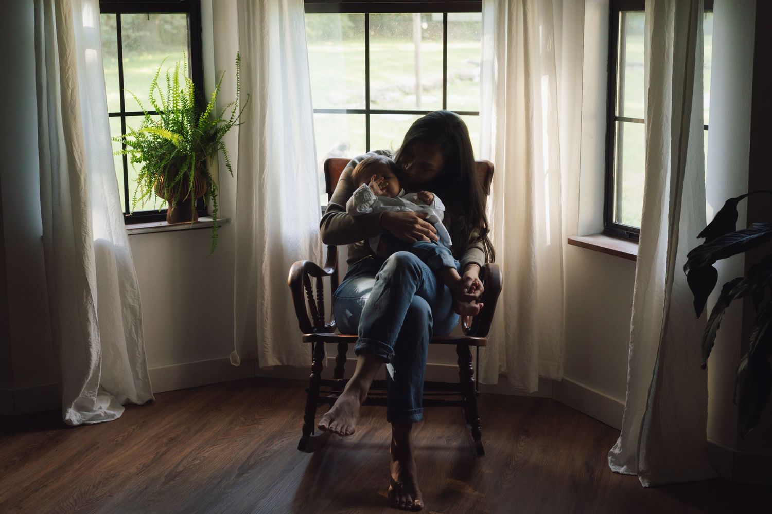 A person in jeans sits in a bright sunlit room with white curtains and potted plants by the window.