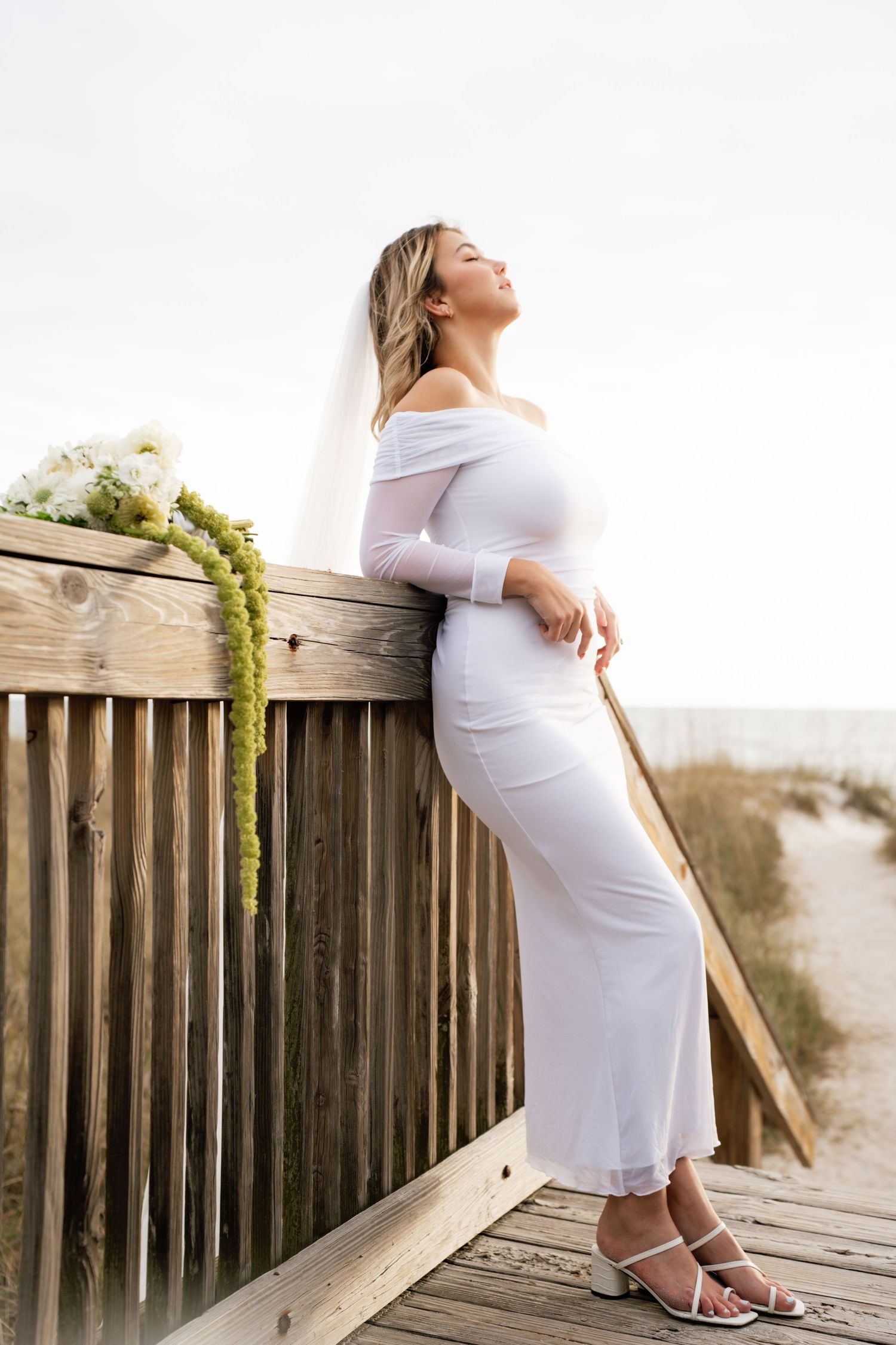 Expectant mother in white dress poses against wooden railing at sunset on beach boardwalk.