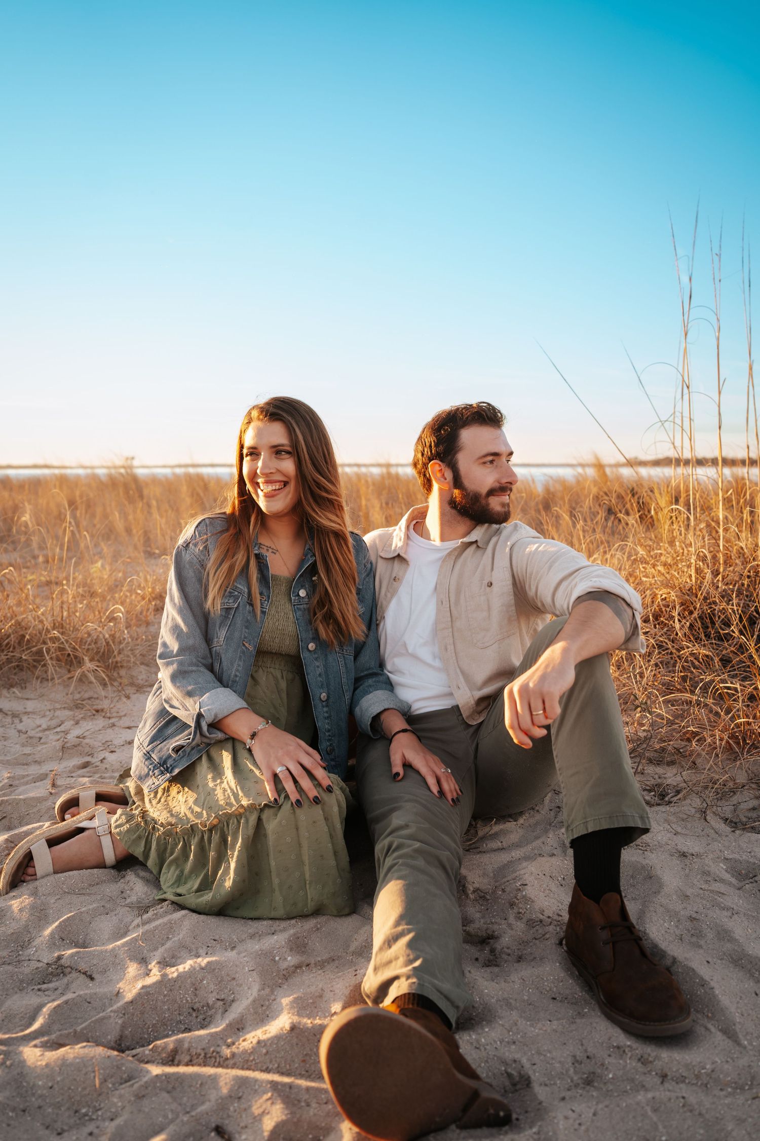 A couple sits together on sandy beach during golden hour with tall grass and blue sky in background.