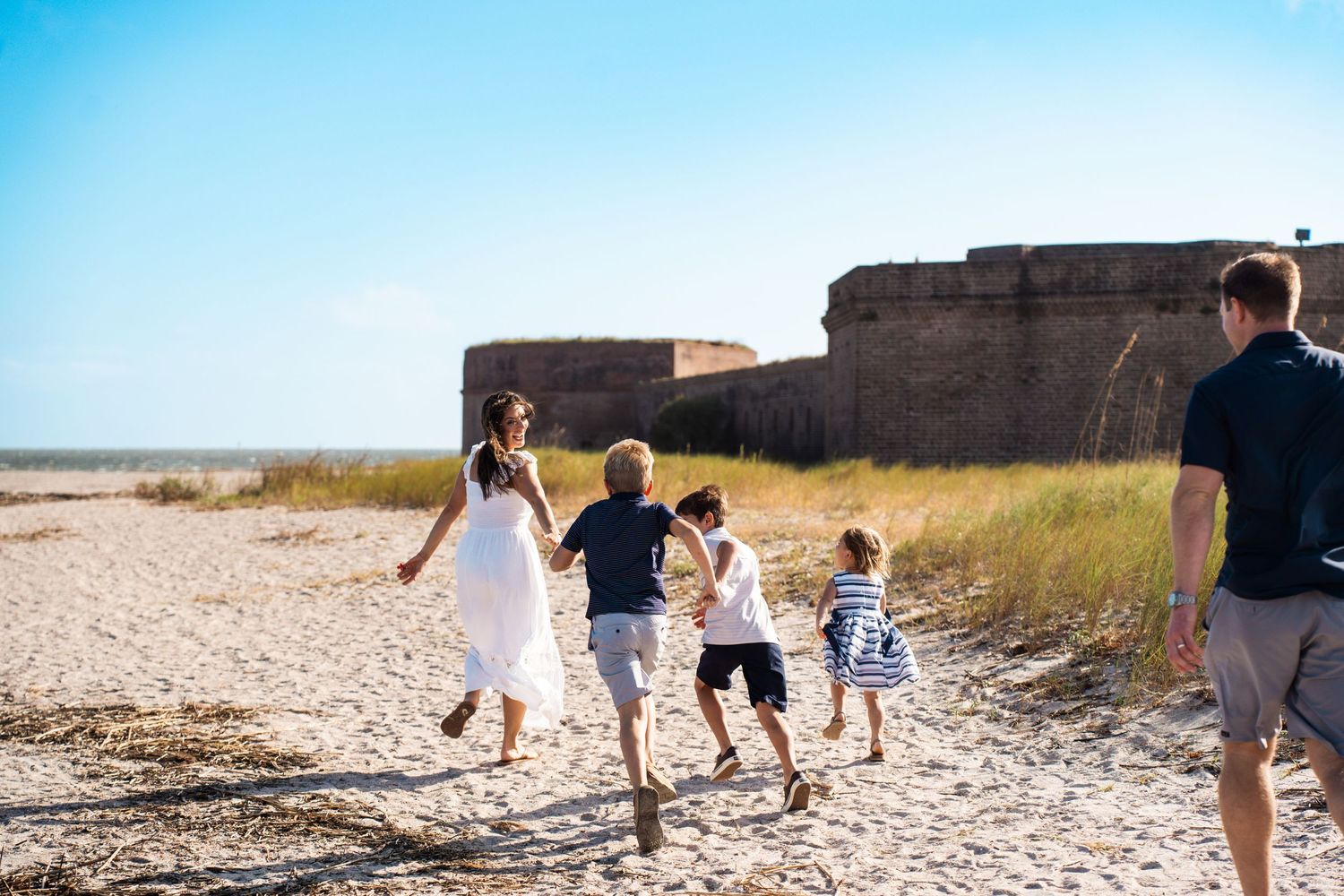 Family running along sandy beach path with historic fort wall in background.