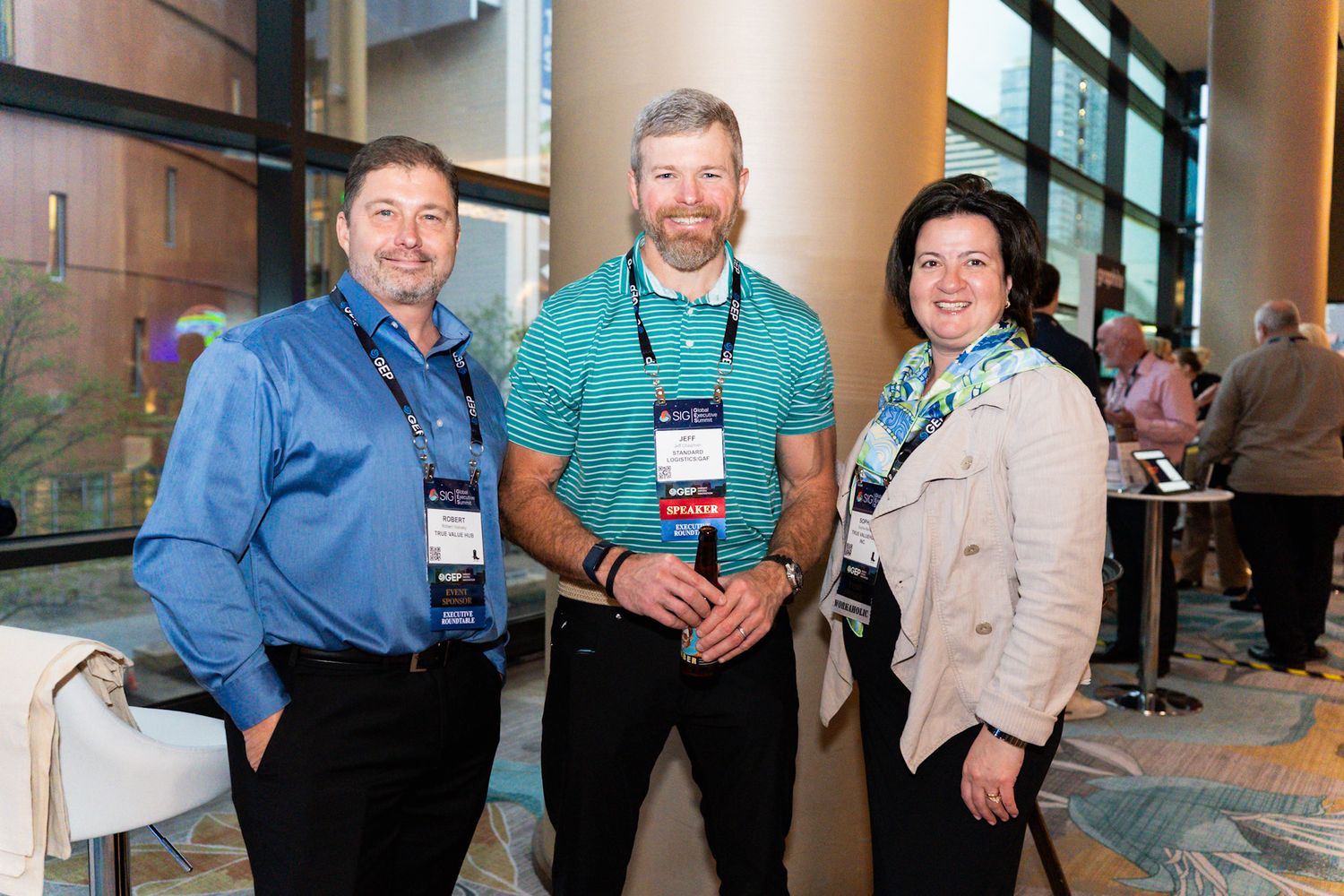 Three professionals wearing conference badges smile together in modern convention center lobby.
