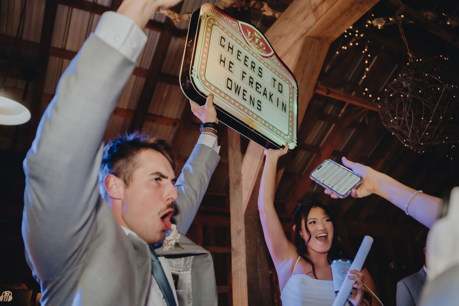 Excited wedding guests holding up a marquee sign at a rustic barn reception celebration.