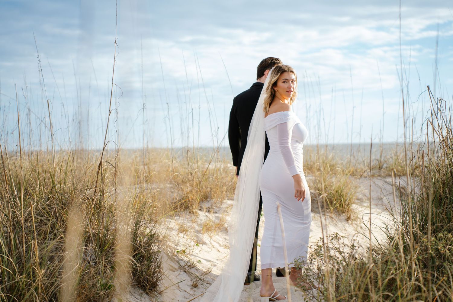 A figure in a white dress walks through tall grass and sand dunes along a coastal beach landscape.
