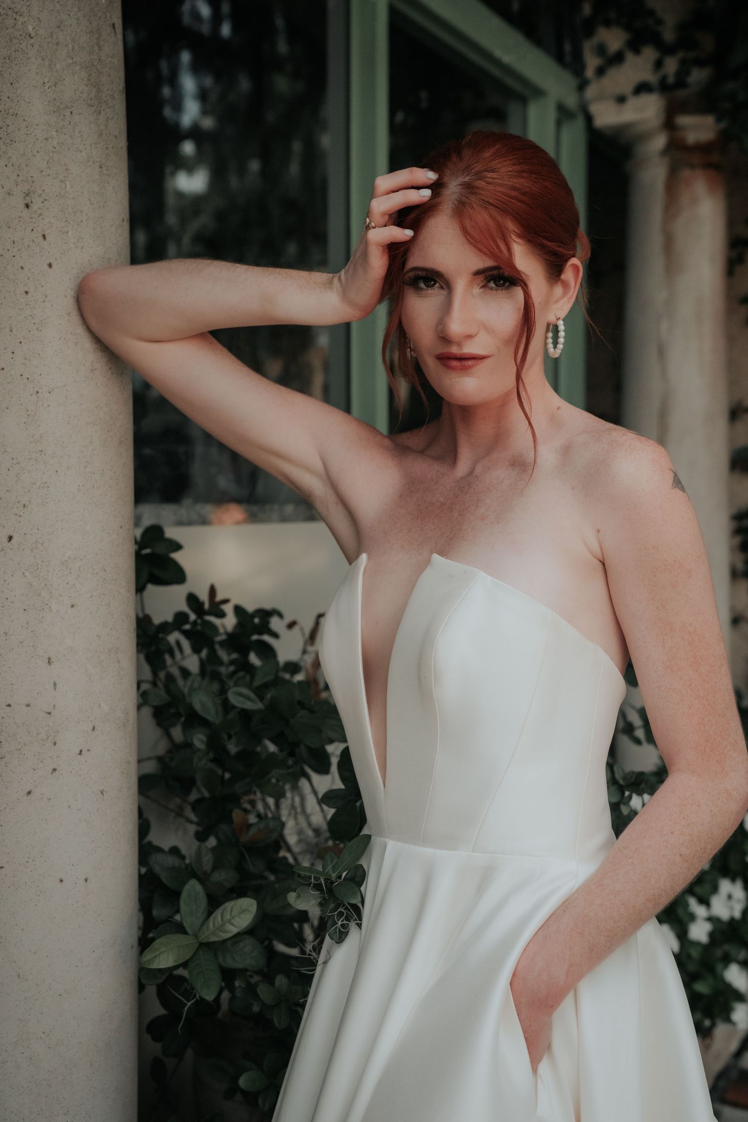 An elegant white strapless wedding dress with clean lines and pockets, styled with red hair and natural foliage backdrop.