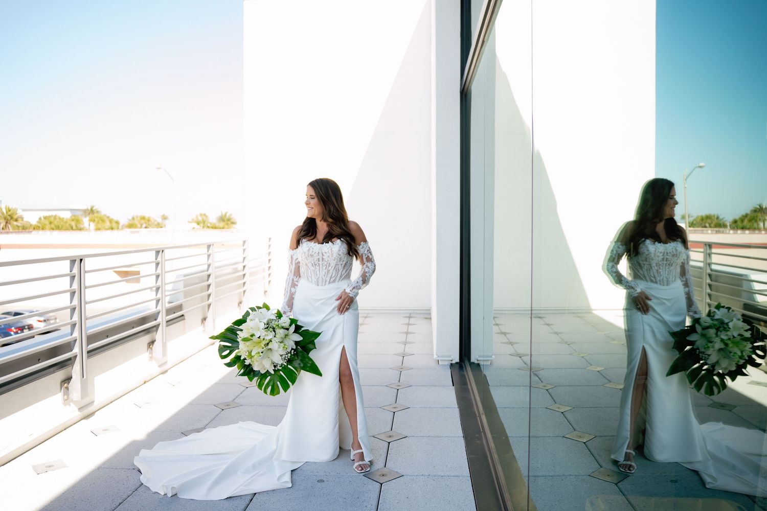 Bride in white gown holding tropical bouquet on modern balcony with reflection in glass wall.