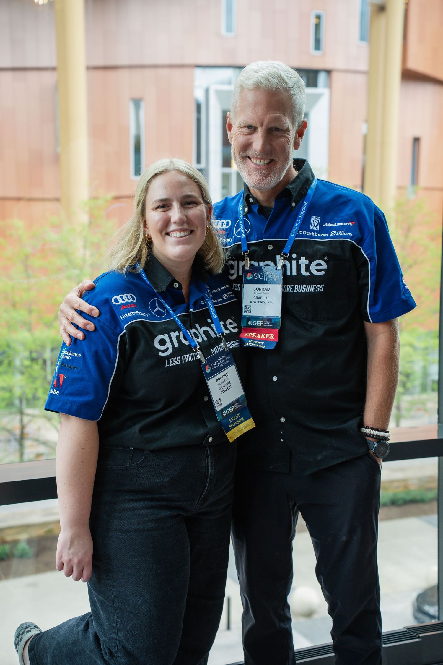 Two people wearing matching Graphite branded blue polo shirts smile together at an indoor corporate event.