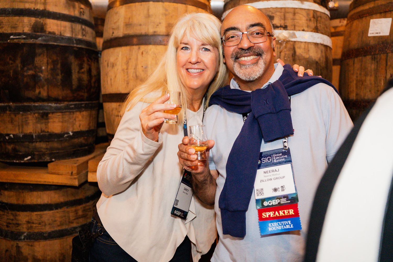 Two smiling attendees holding drinks pose among wooden barrels at a speaker conference event.