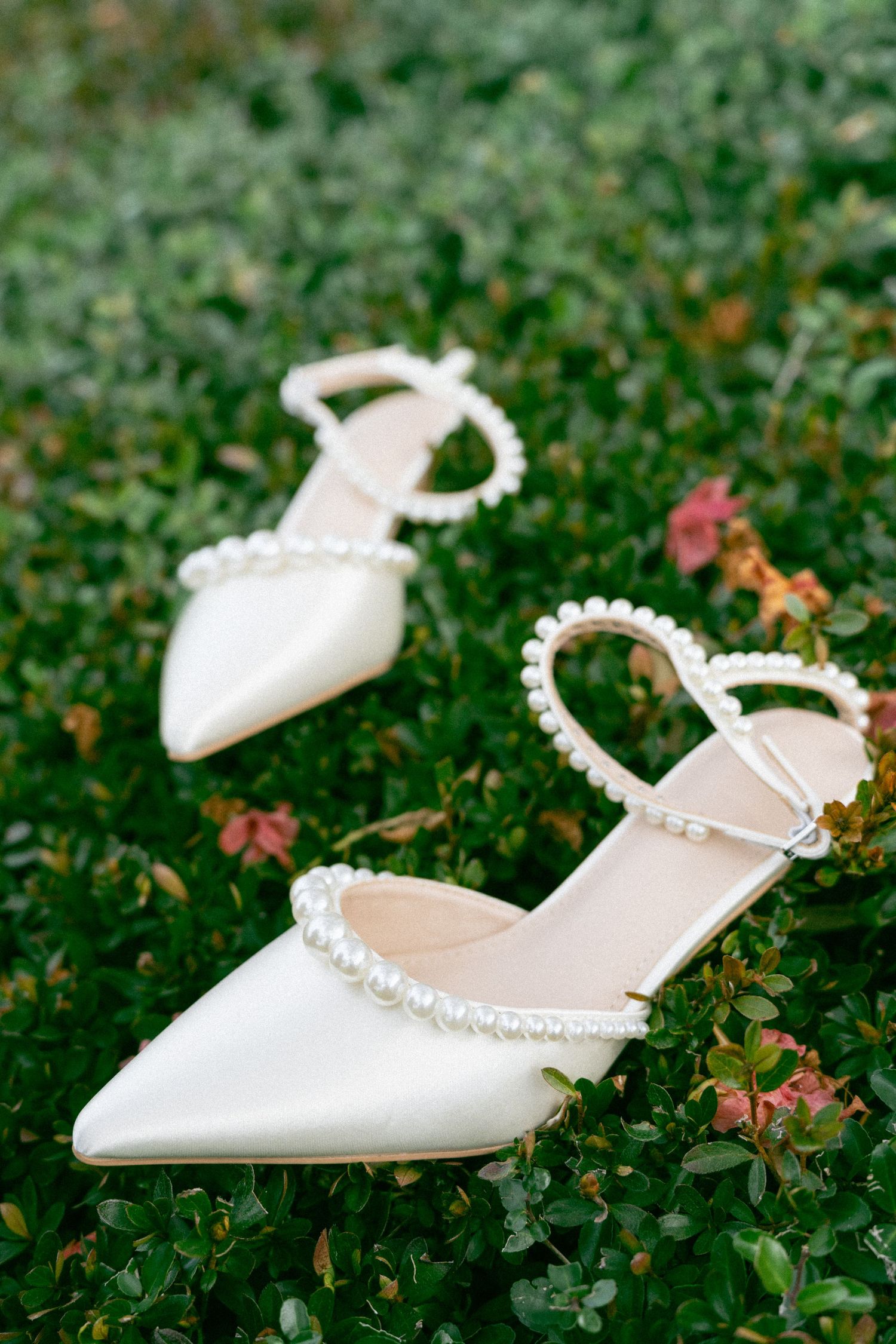 Elegant white bridal slingback heels with pearl straps photographed laying on green grass and pink flowers.