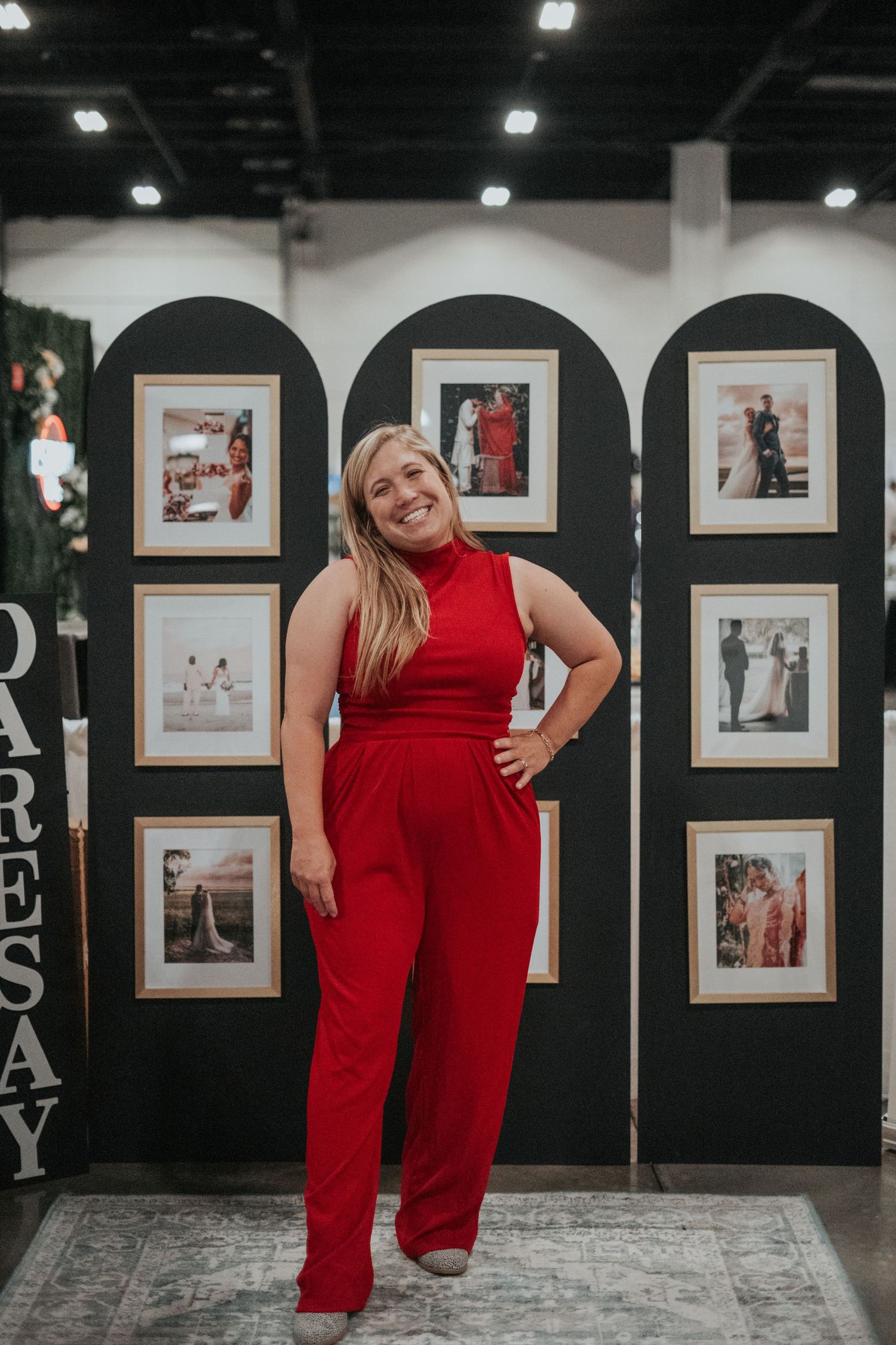 Woman in a vibrant red jumpsuit poses confidently in front of a wall display of framed photographs at Premier Bride Expo in Jacksonville, FL