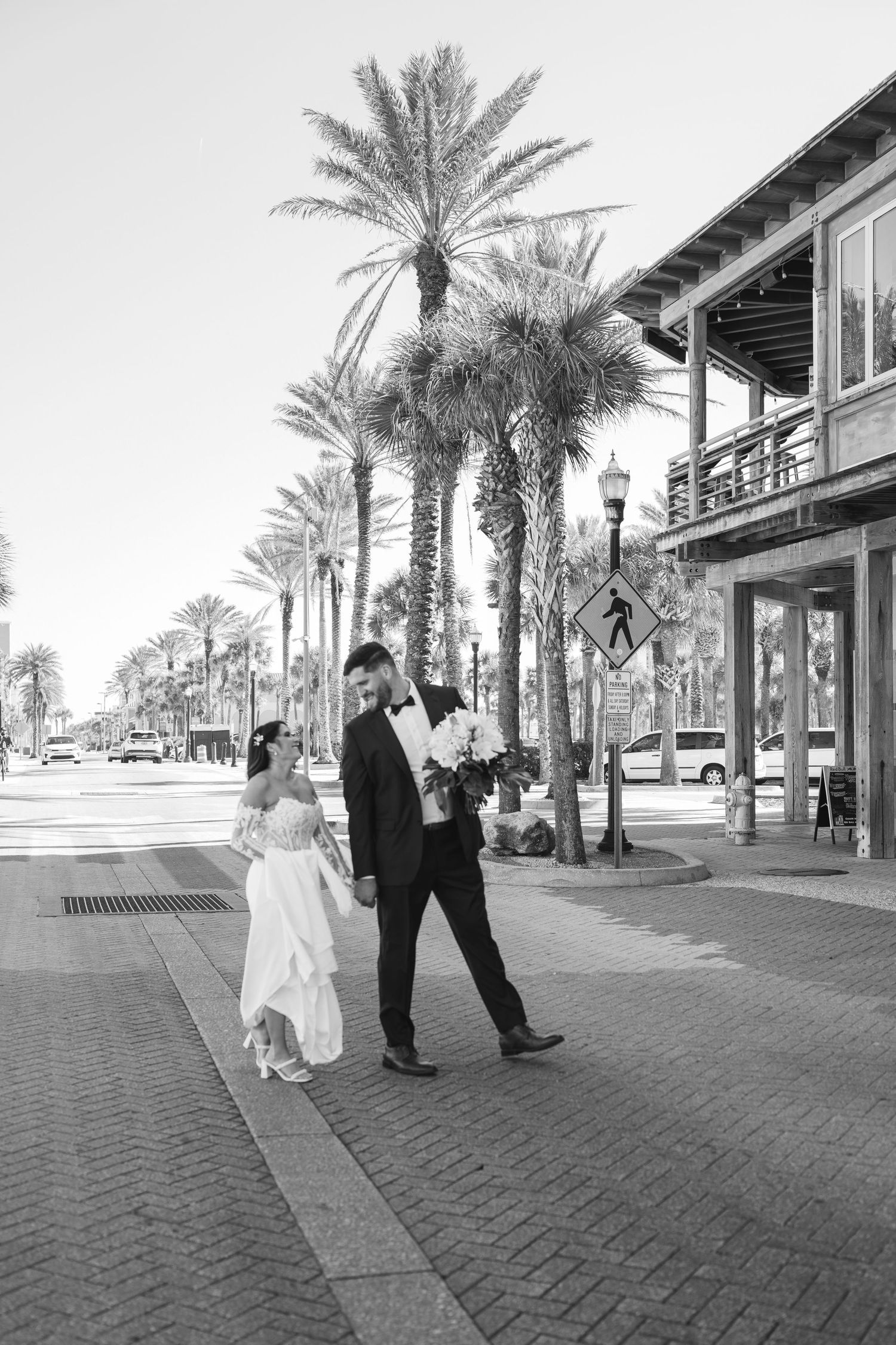Black and white photo of a bride and groom walking hand in hand on a palm tree-lined sidewalk on their wedding day.