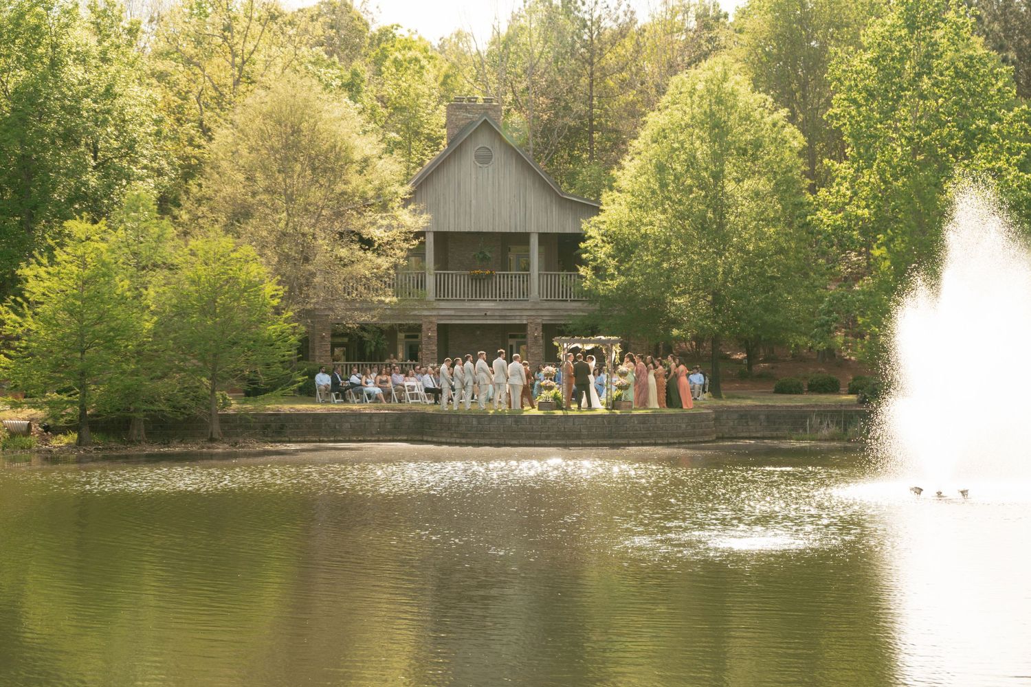 Outdoor wedding ceremony by a serene pond with a rustic two-story cabin surrounded by lush green trees.
