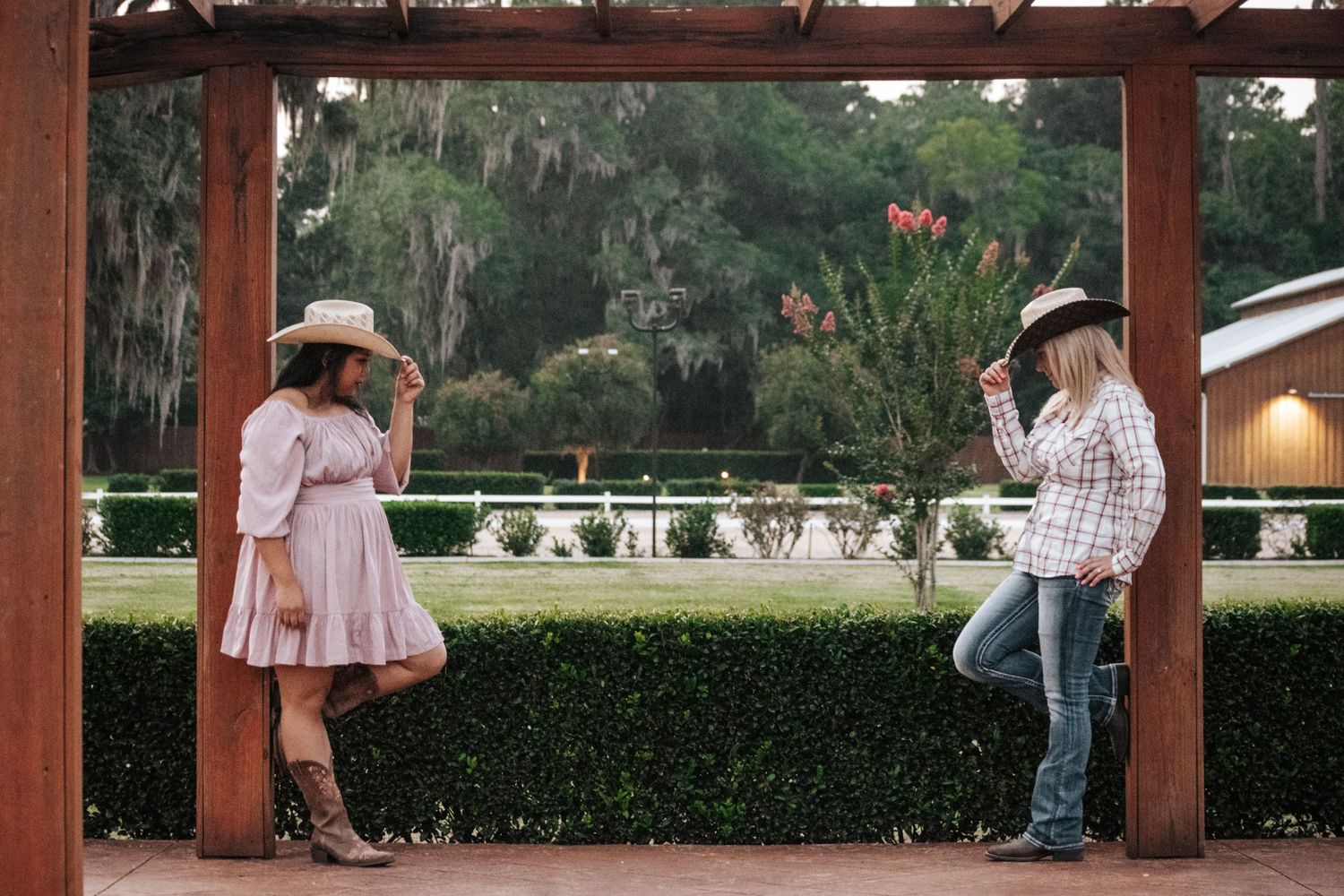 Two people in western attire and hats stand beneath a wooden beam structure in a garden setting.
