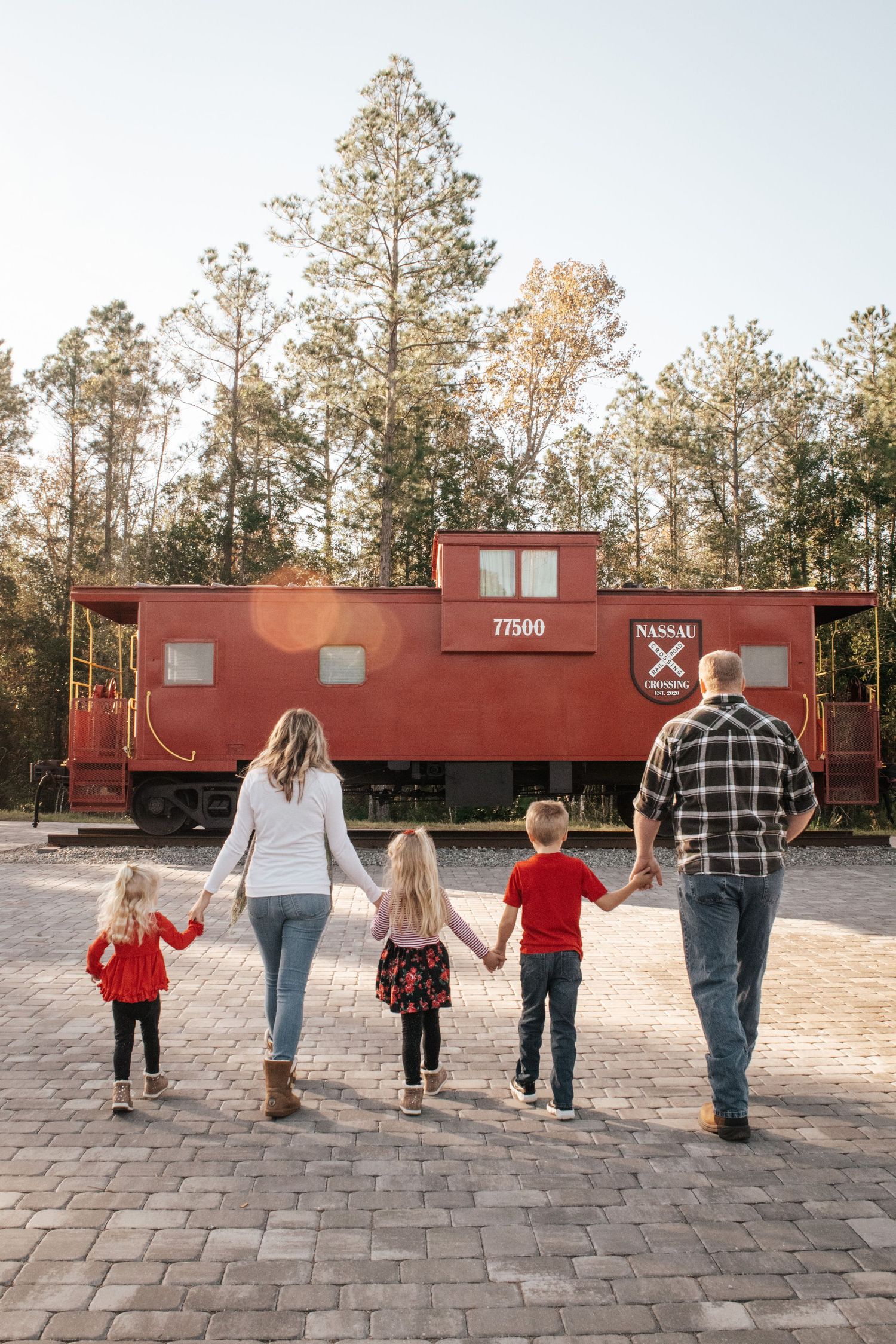 Family walking hand in hand toward a vintage red caboose in a pine forest setting.