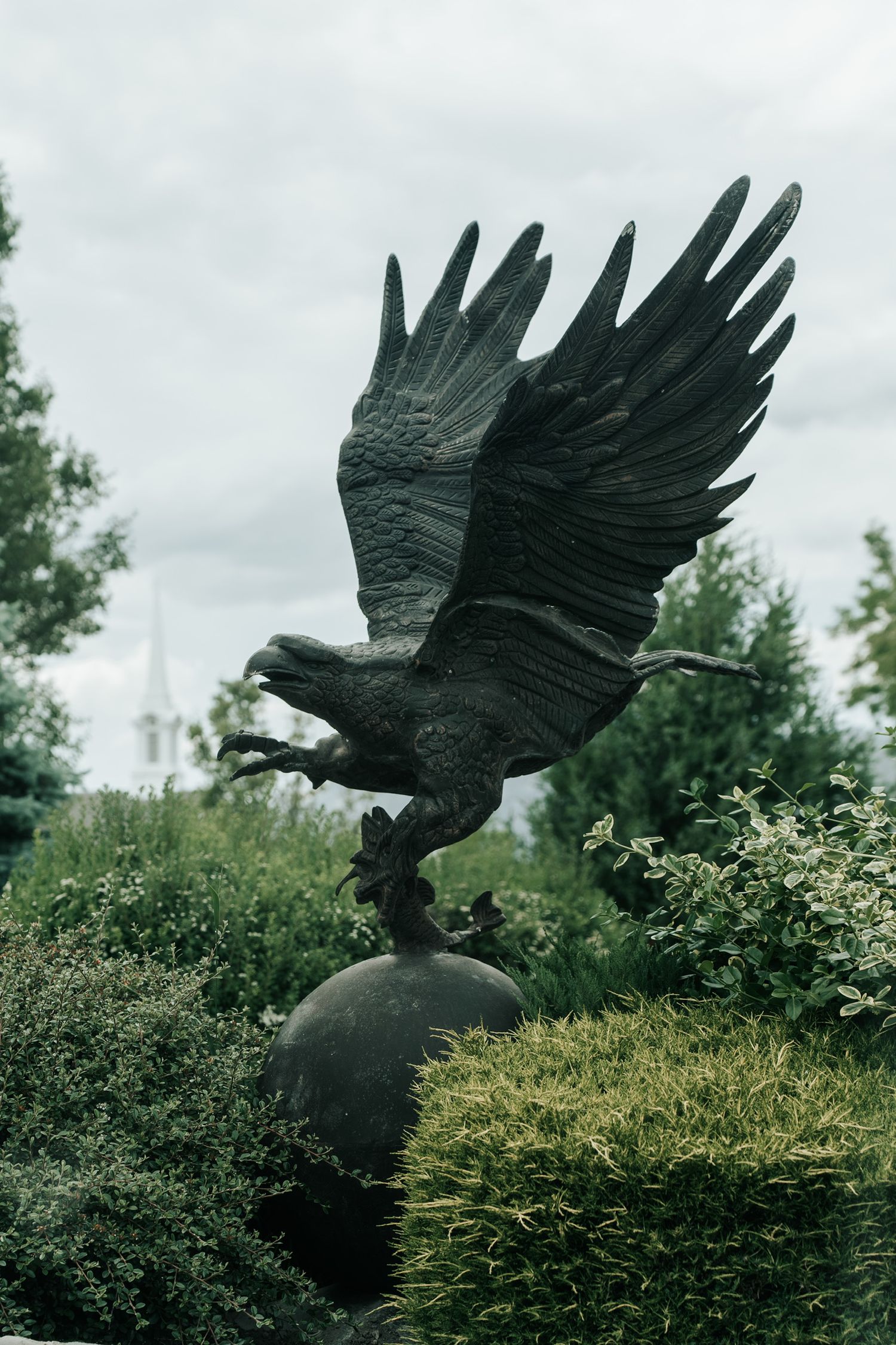A large bronze eagle statue with spread wings perches atop a spherical base in a garden setting.