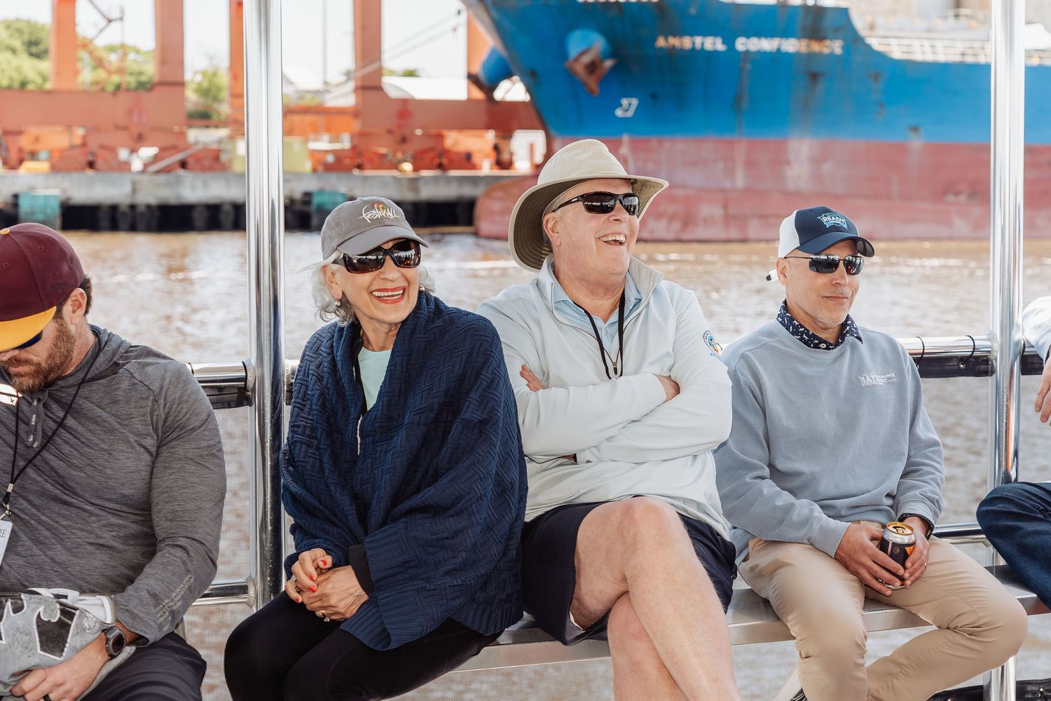 Group of smiling adults in sunglasses seated outdoors near a large cargo ship.