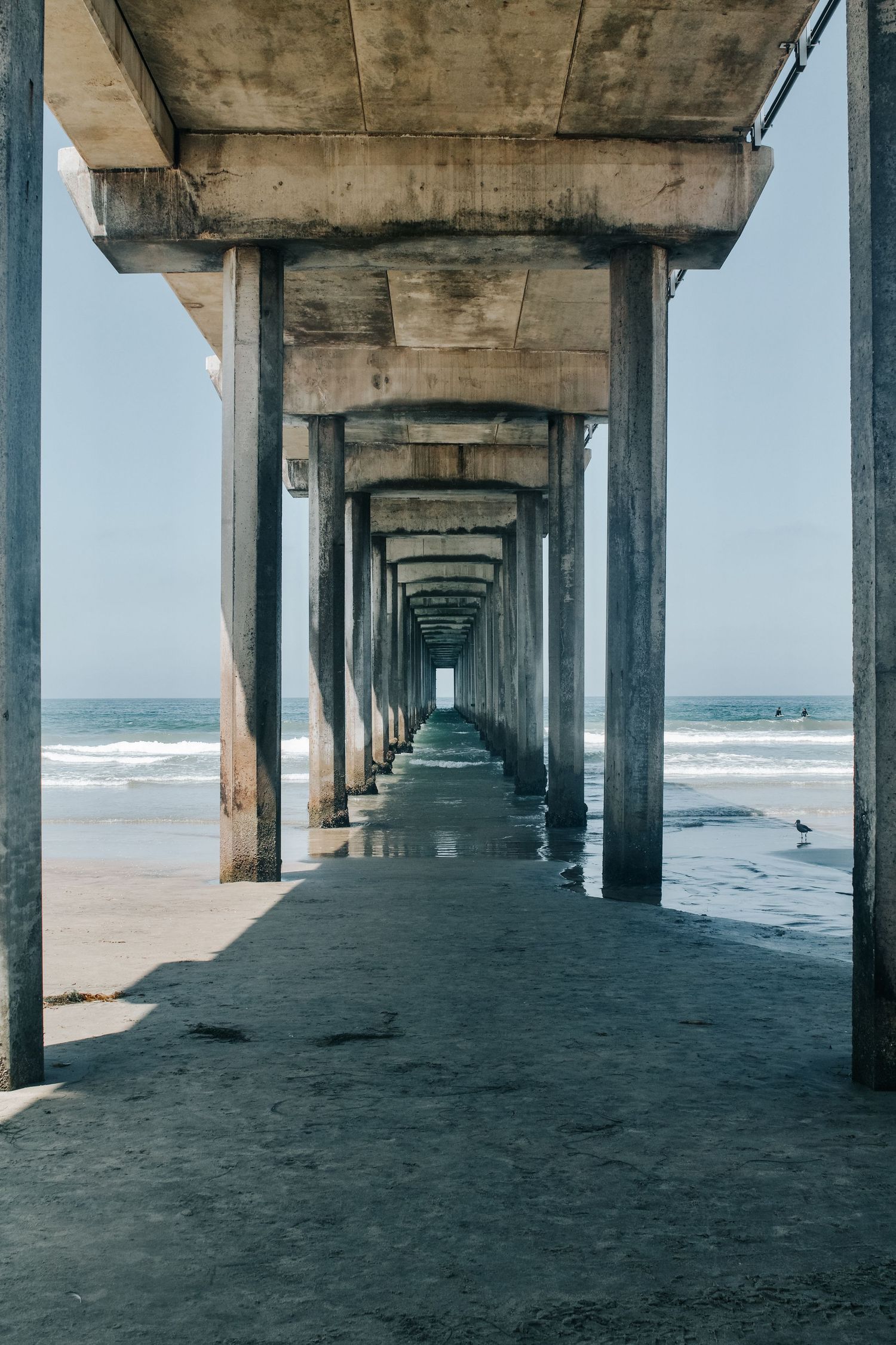 View under a concrete pier stretches toward the ocean horizon with waves crashing on the beach.