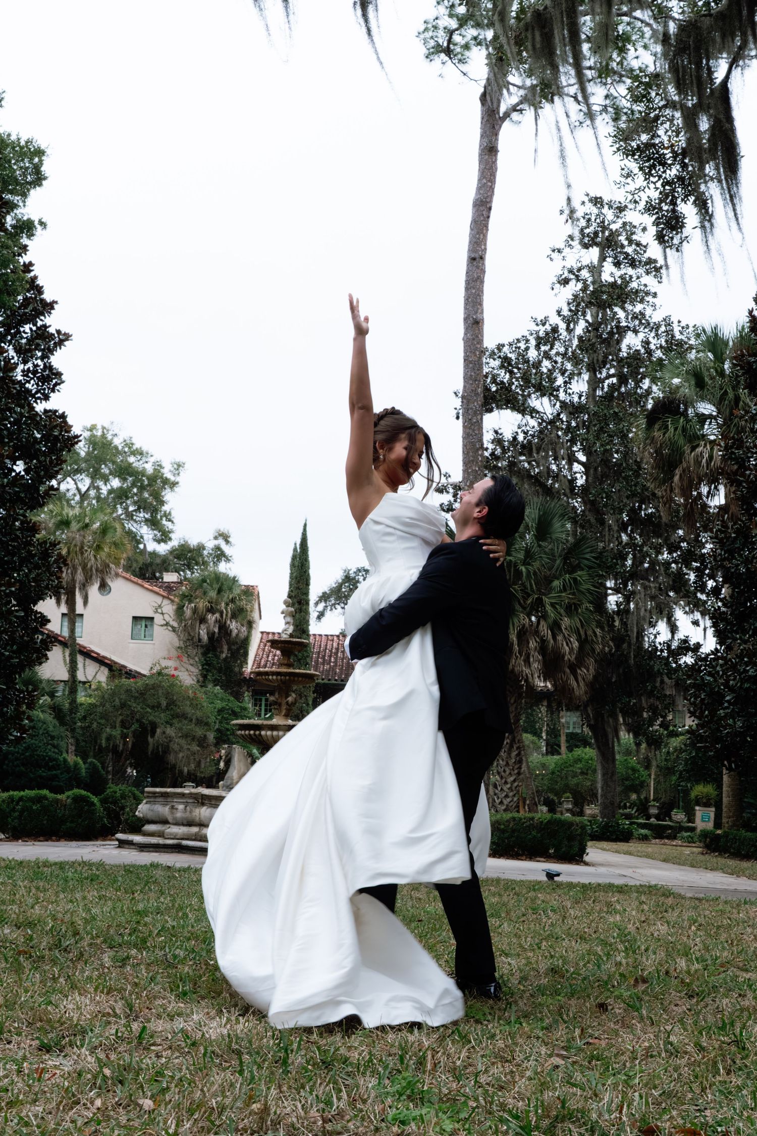 A couple in formal attire performs a romantic dip pose outdoors with trees and greenery in the background.