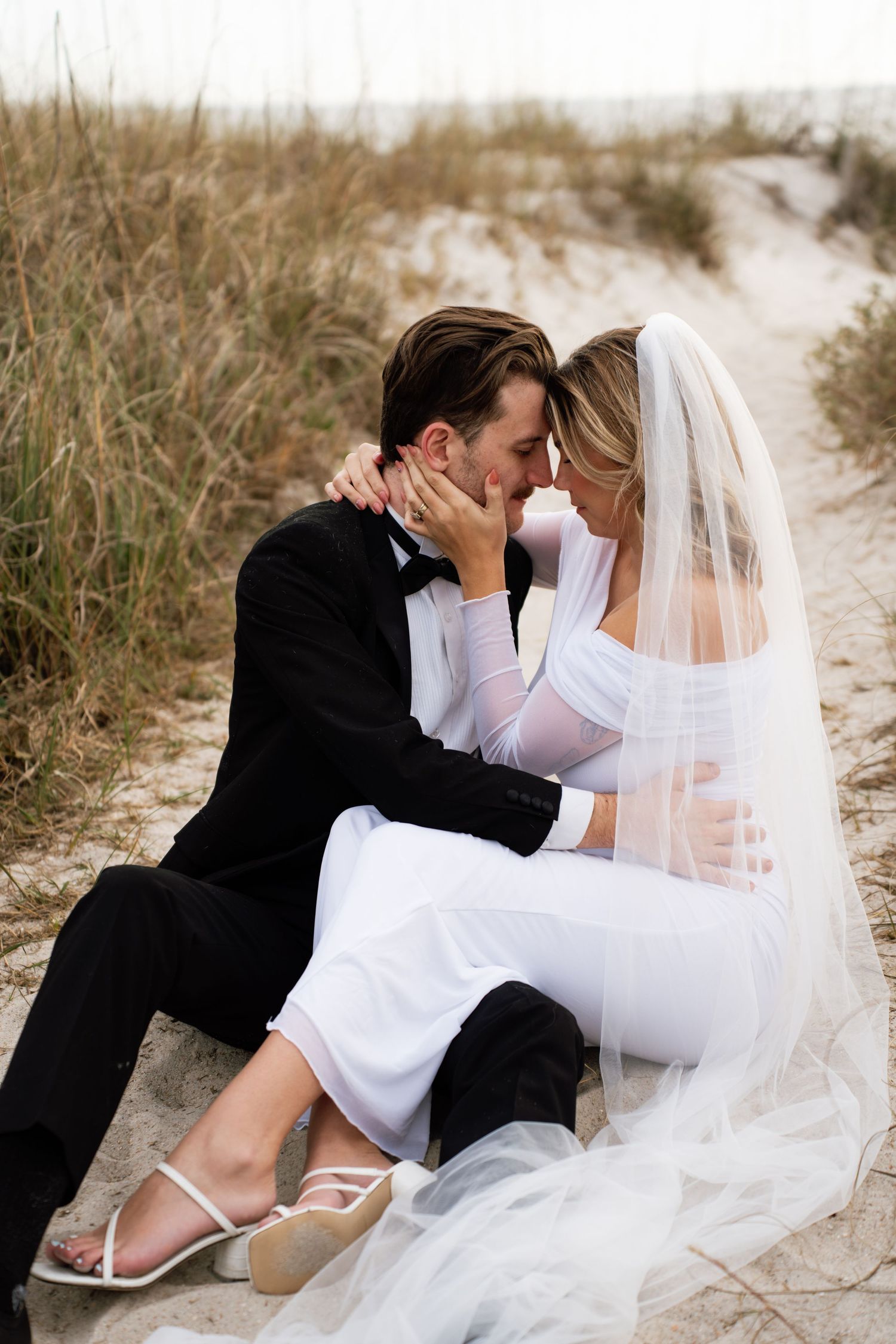A romantic beach wedding photo series showing a couple in white and black attire embracing on sandy dunes.