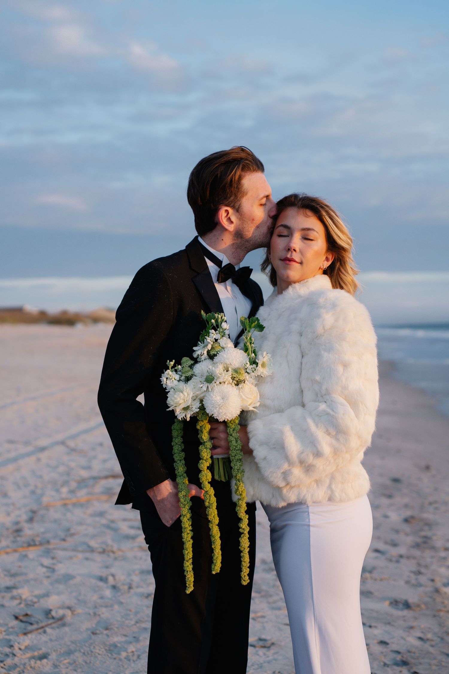 A couple shares romantic moments on a sandy beach at sunset wearing formal black and white wedding attire.