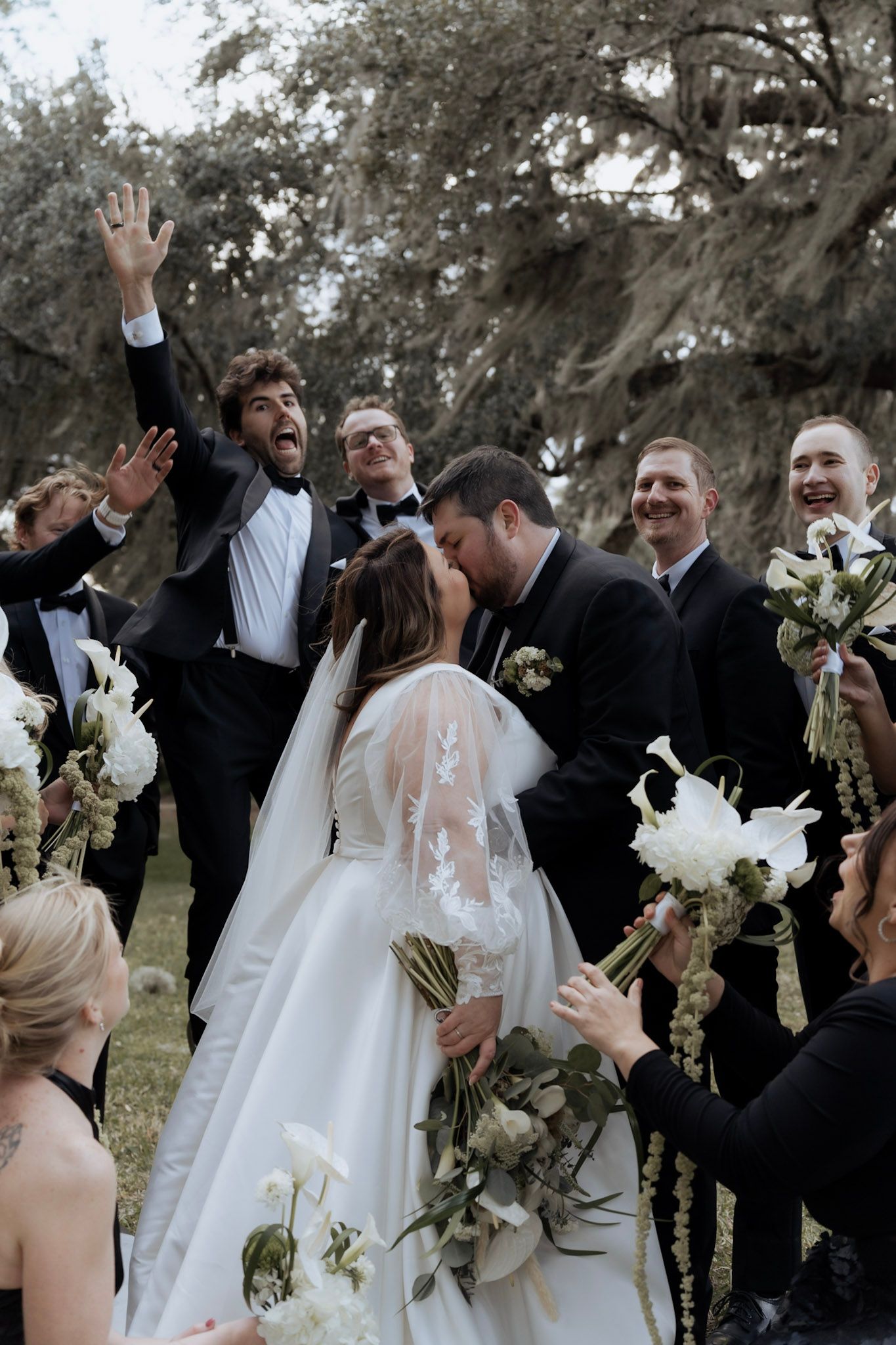 Wedding party celebrates joyfully outdoors with raised hands and bouquets during group photo session.
