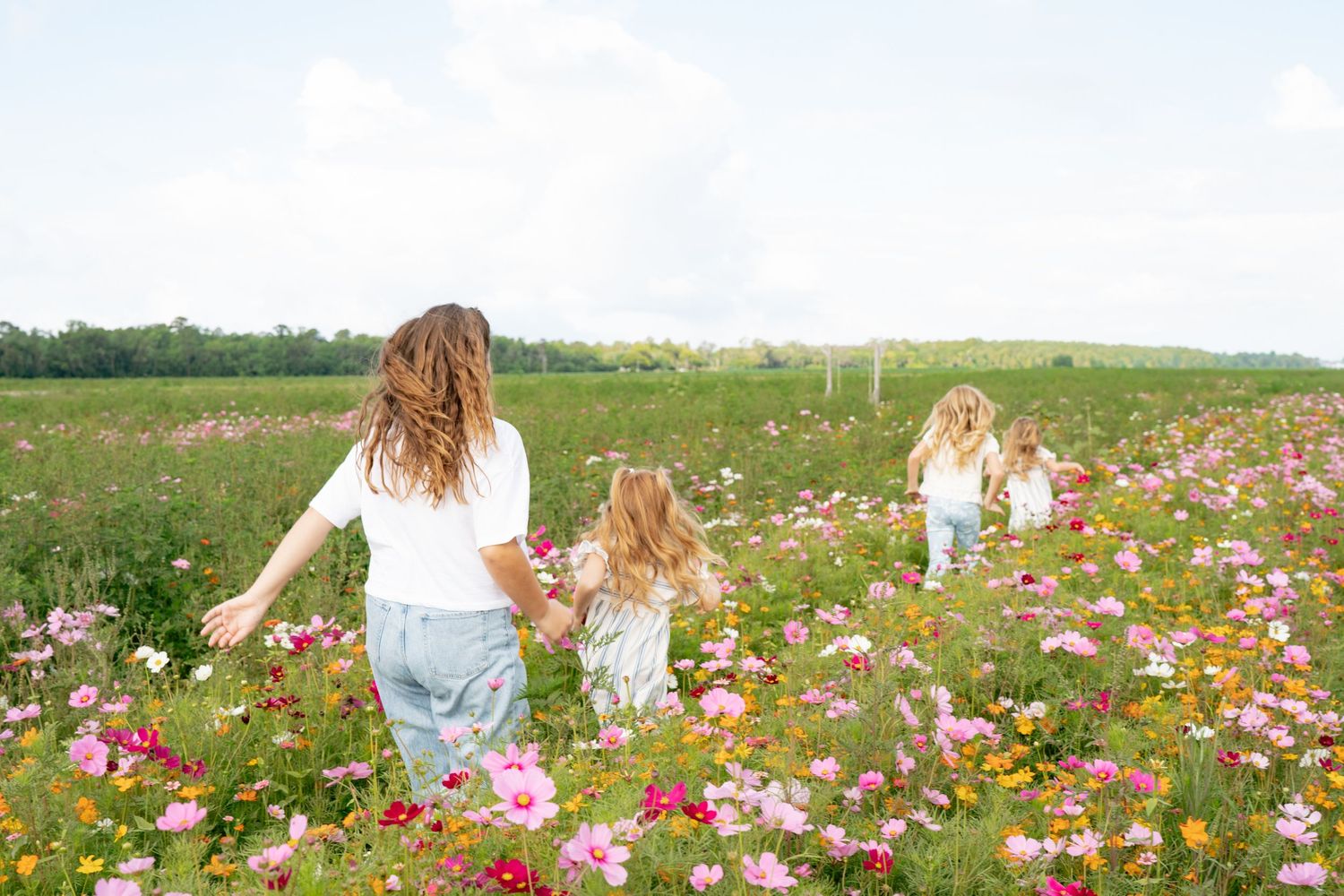 Three females walk through a vibrant wildflower field with pink and white blooms under a bright cloudy sky.