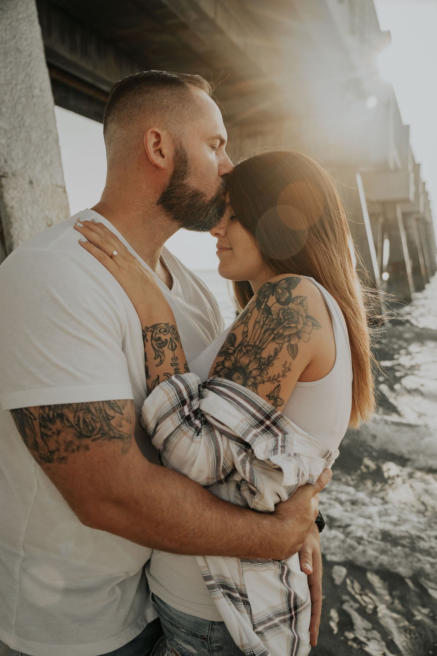 A romantic couple with tattoos embrace under a pier during golden hour as sun flares illuminate their tender moment.